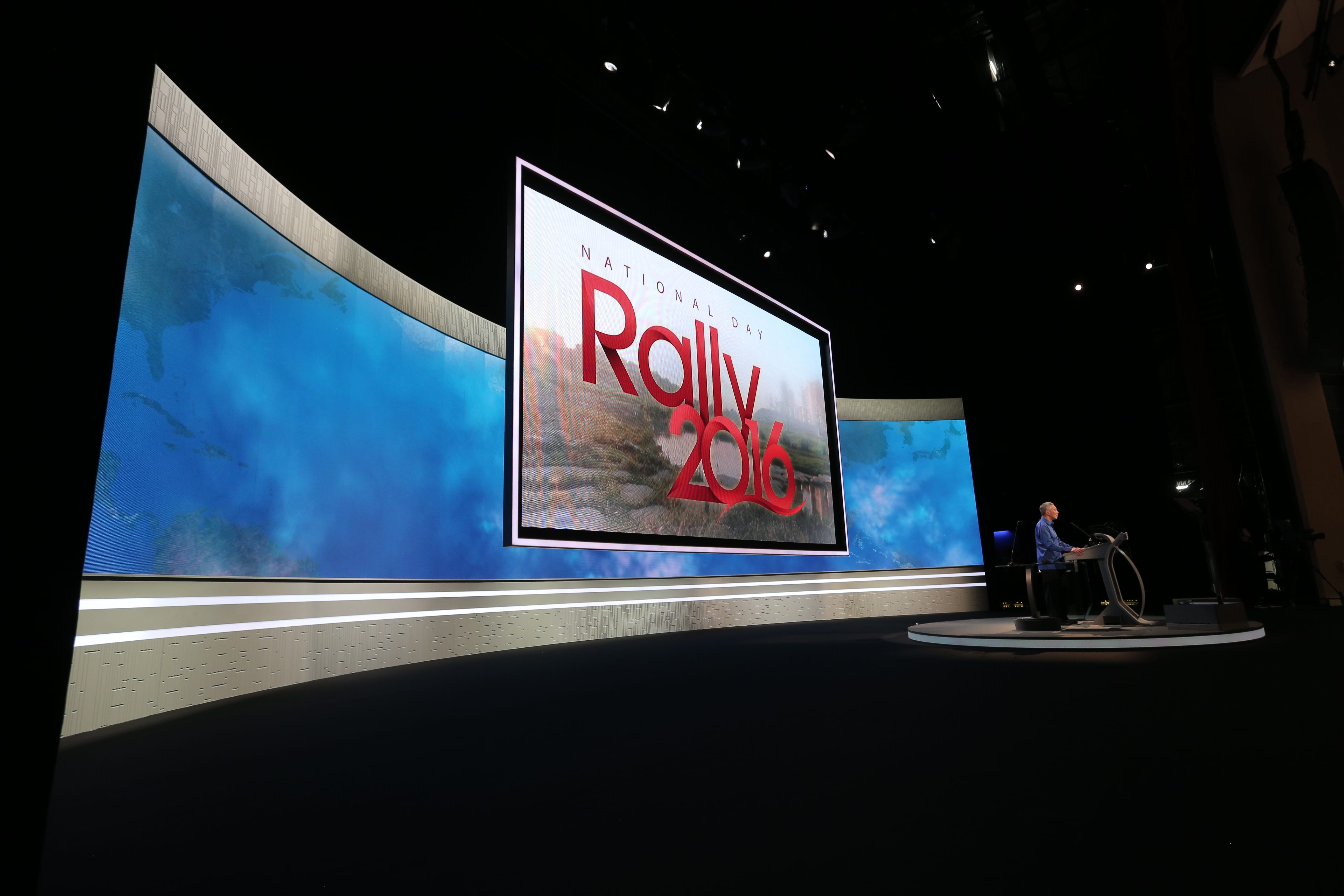 Stage set with large screen displaying "National Day Rally 2016," speaker at podium.