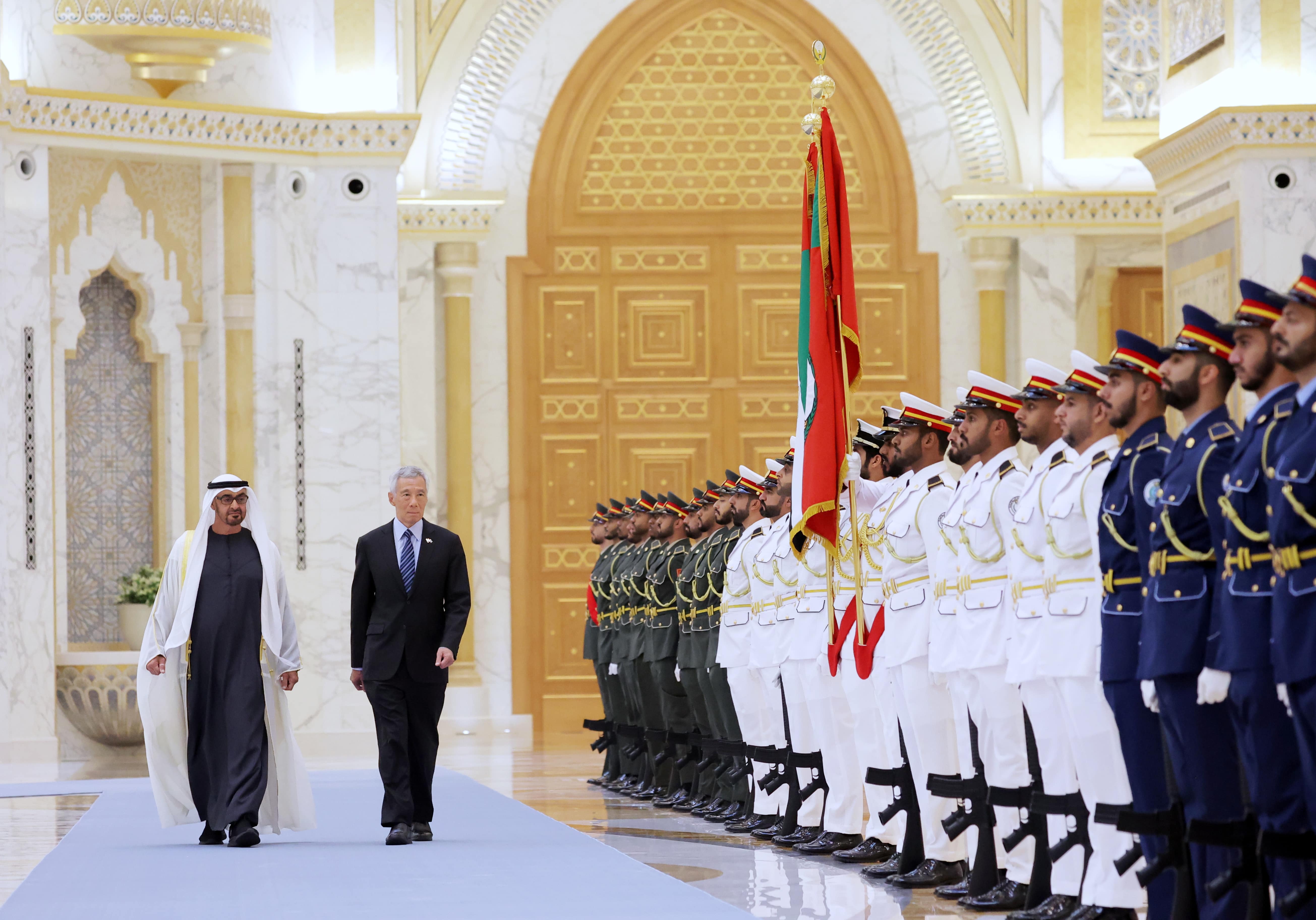 Two men walk past a military honor guard in ornate hall with the UAE flag.