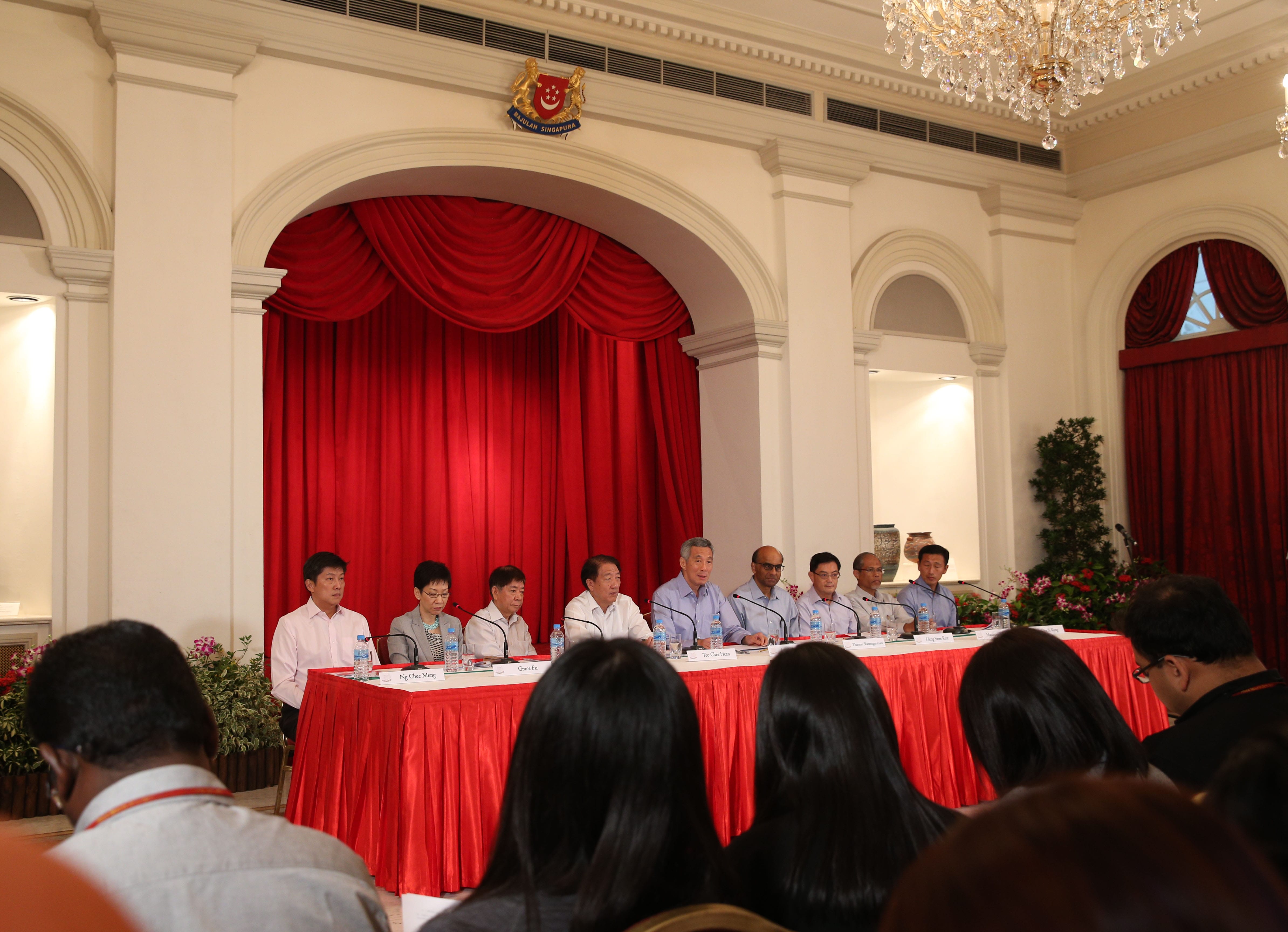 Panel of people seated at a table with microphones in front of a red backdrop, audience in foreground.