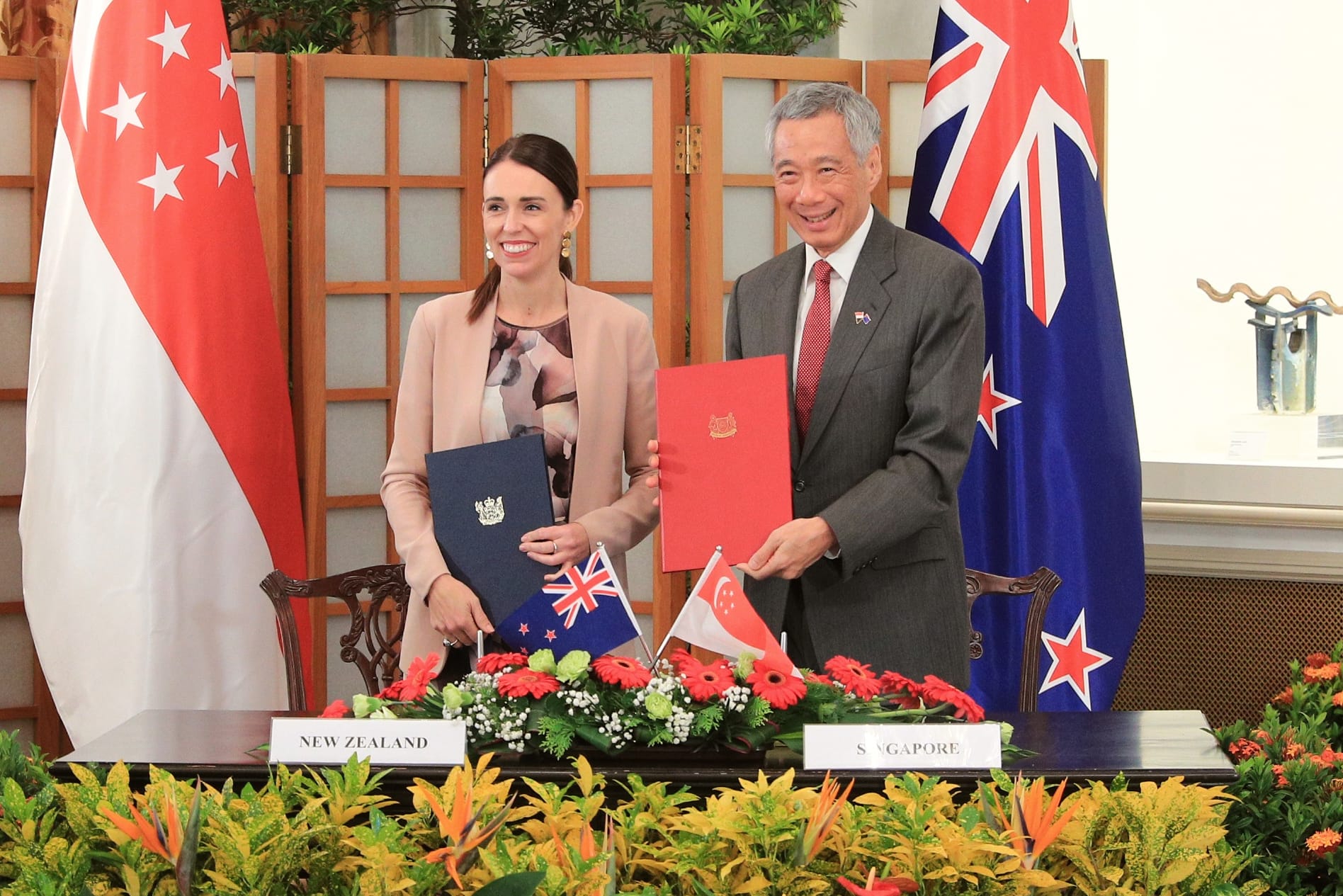 Jacinda Ardern and Lee Hsien Loong hold documents by New Zealand and Singapore flags.