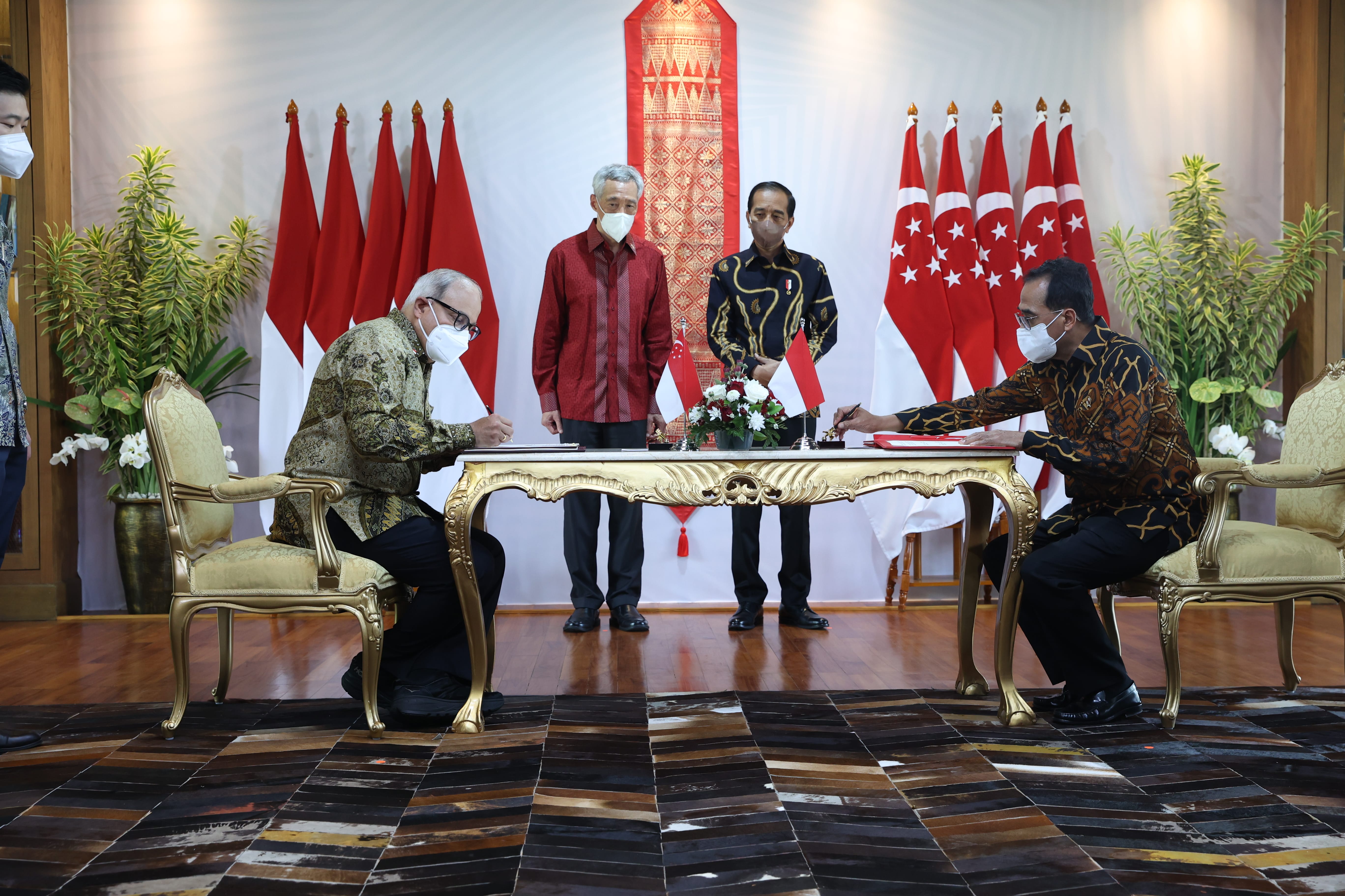 Two people signing documents at a table, Indonesia and Singapore flags behind them, with Jokowi standing in the middle.
