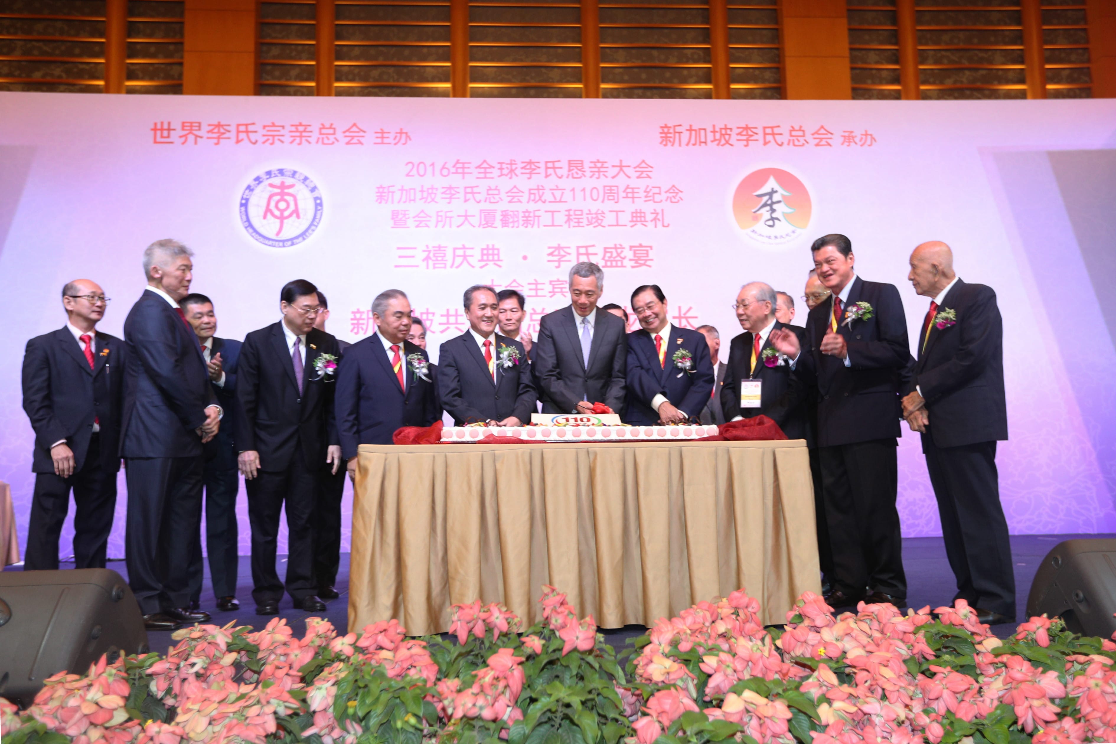 Group of men in suits at a table with a cake, flowers in the foreground.