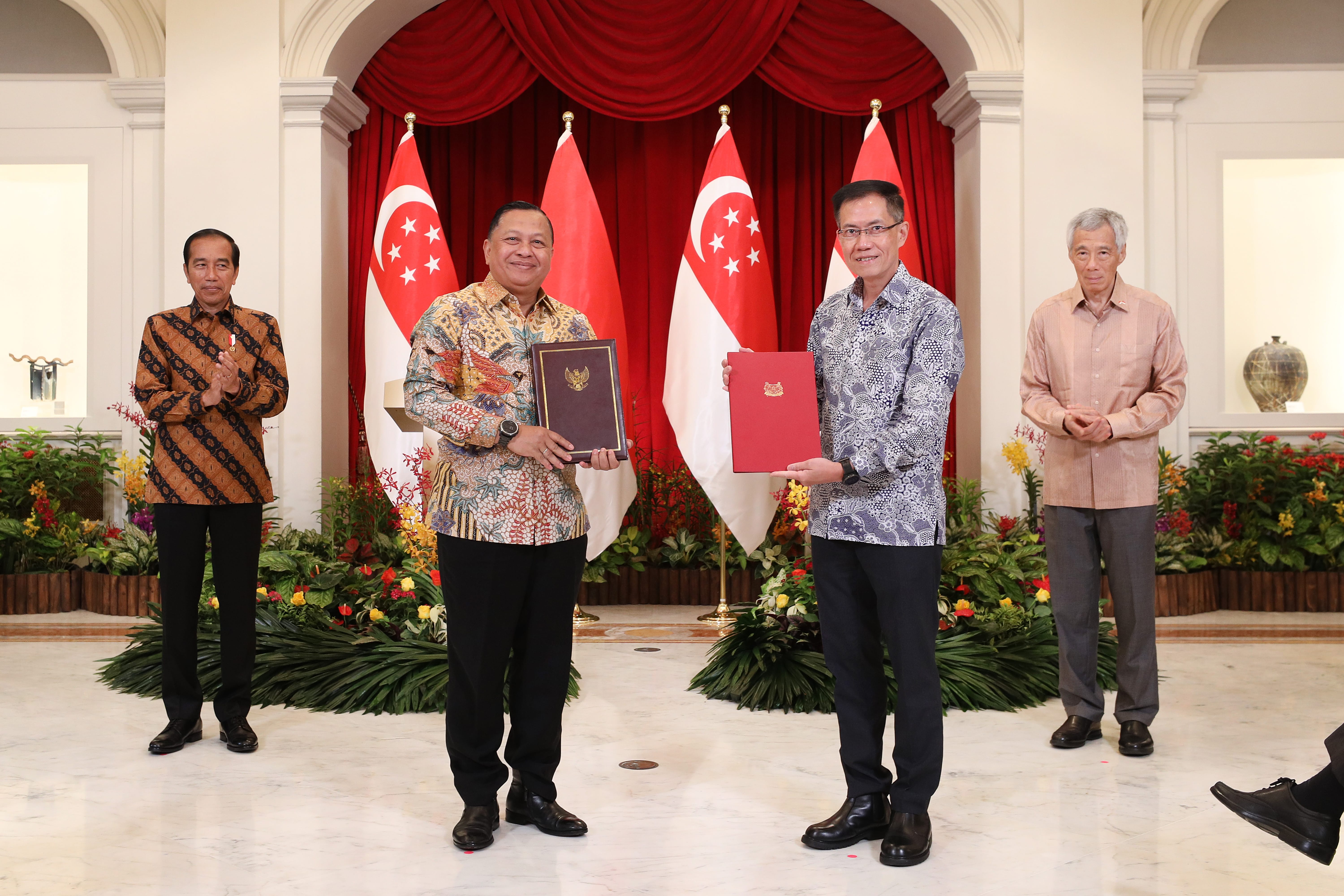 Four men in formal attire holding documents, standing before Singapore flags.