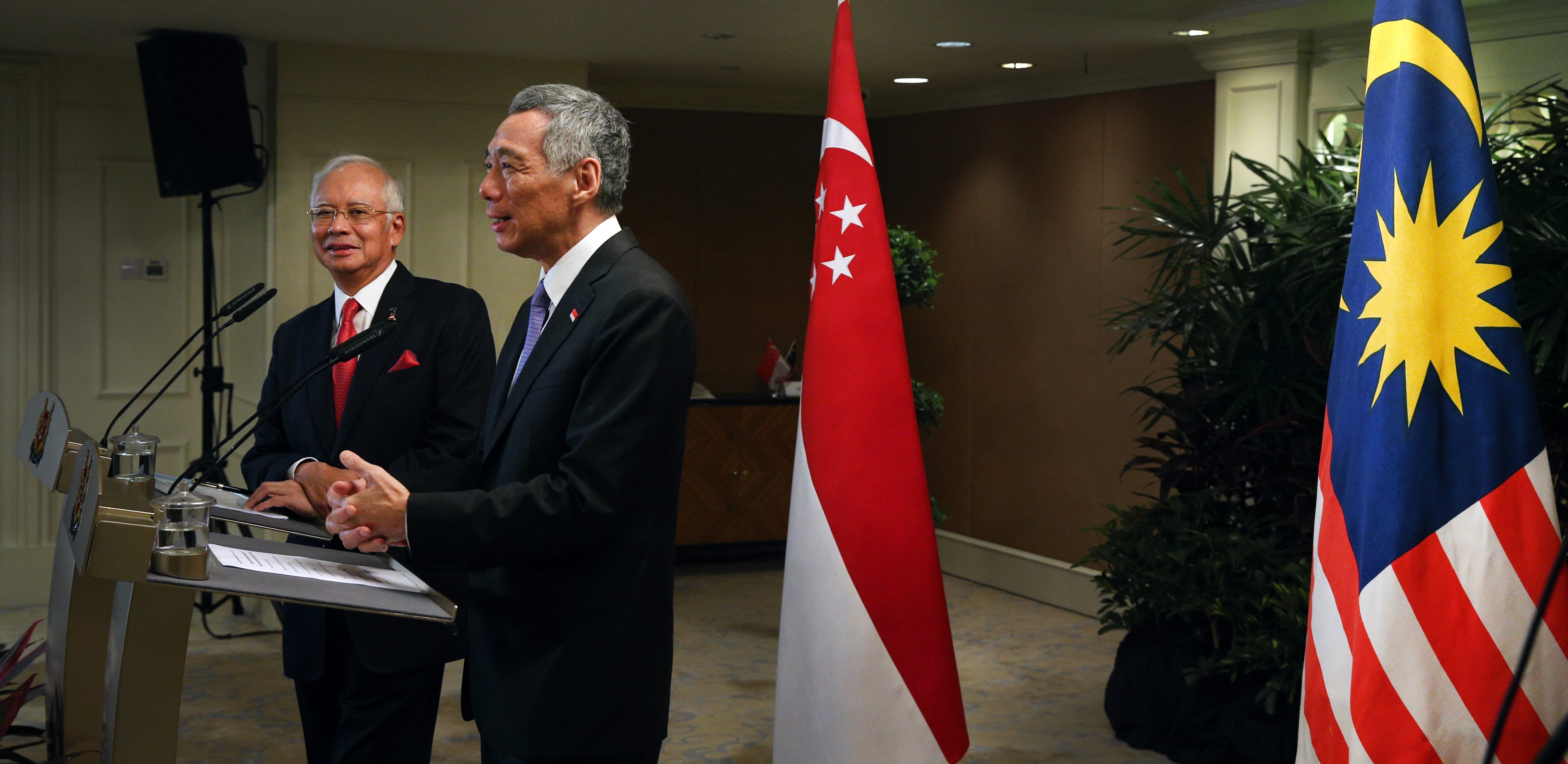 Two men at podiums with microphones, near Singapore and Malaysia flags.