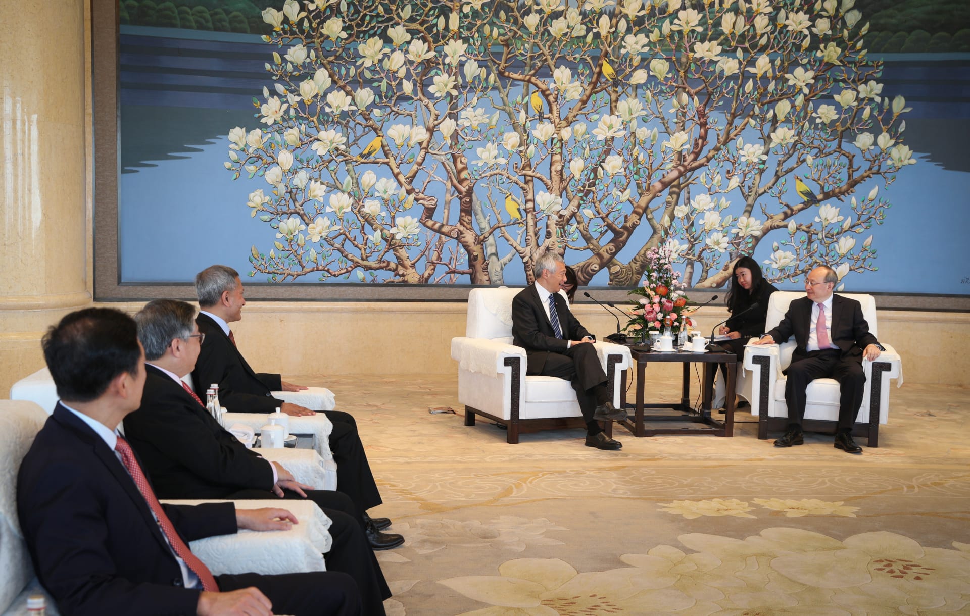 People in suits sit in white armchairs, with a large painting of a flowering tree behind them.