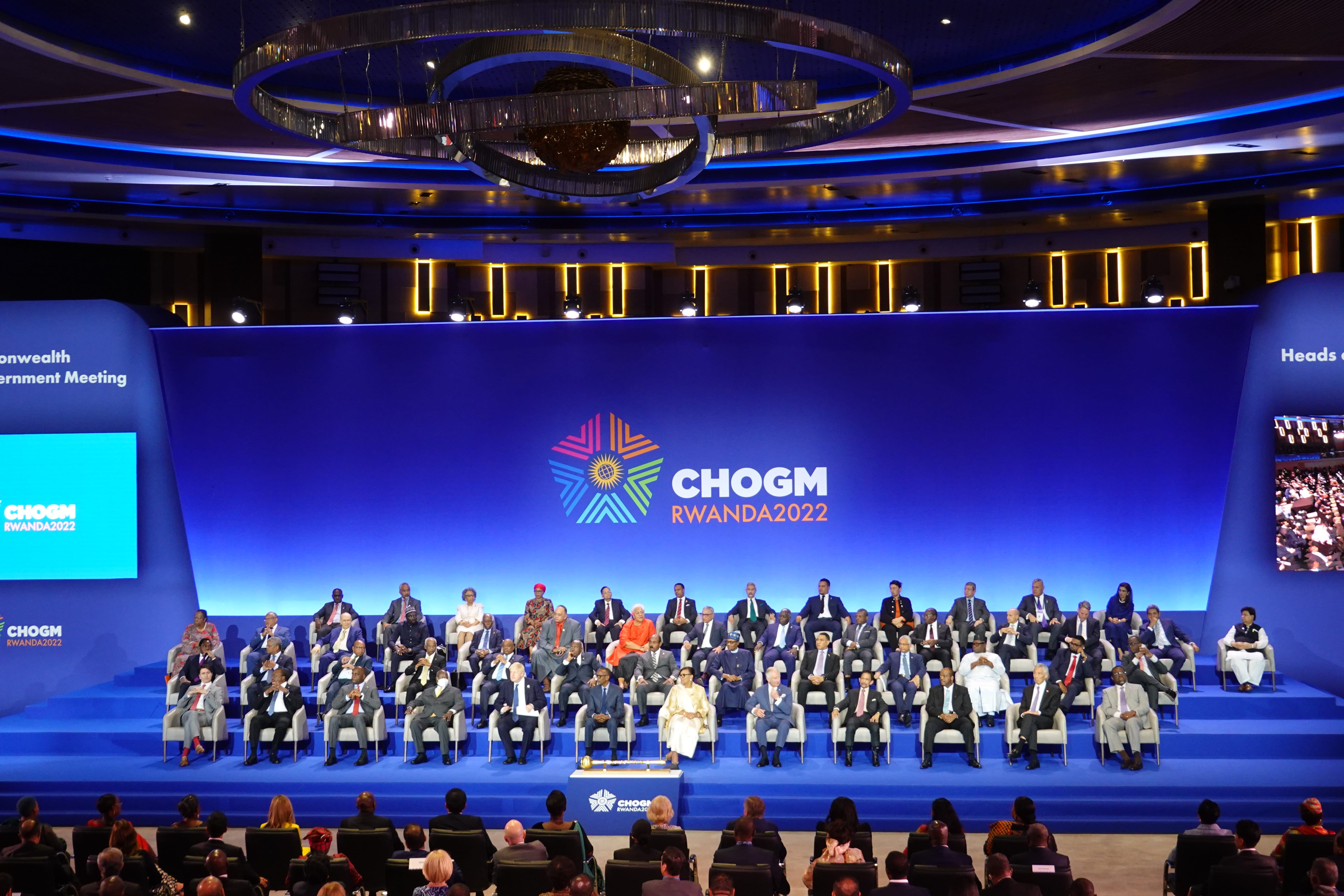 CHOGM RWANDA2022 event with diverse group of people seated on stage and in audience. Blue background.