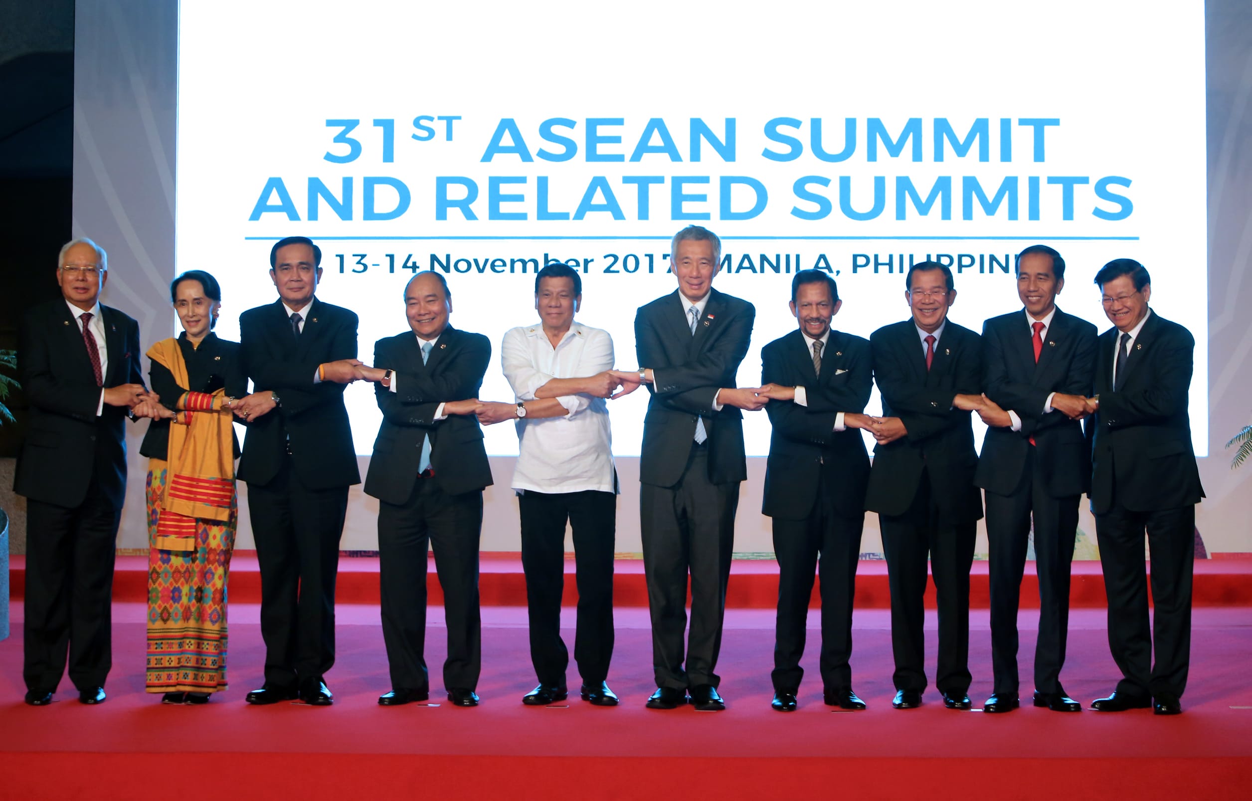Ten people holding hands on red carpet, in front of a screen: 31st ASEAN Summit.