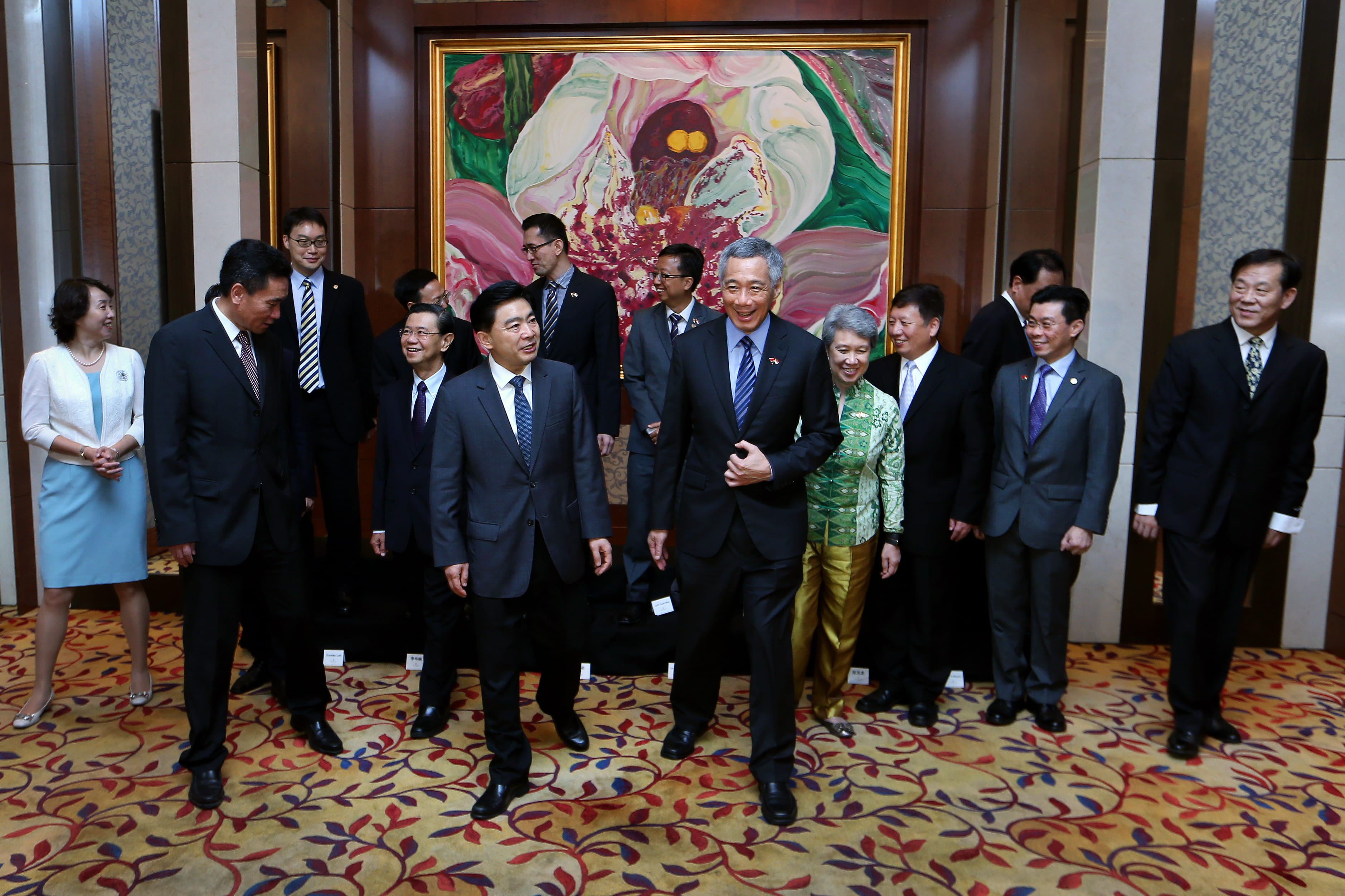 Lee Hsien Loong and others in suits walk, stand, beneath a framed flower painting.