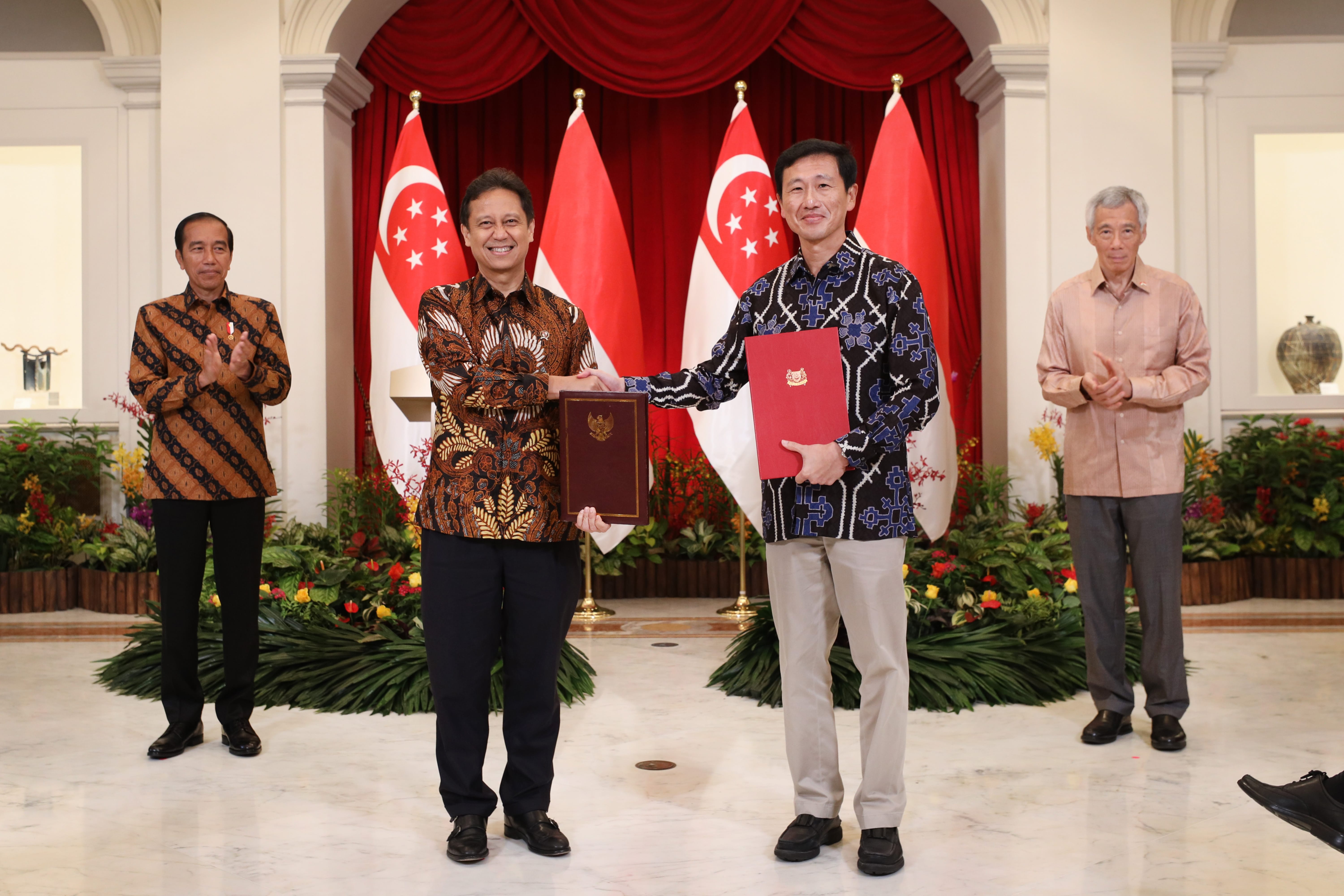 Four men stand before Singapore/Indonesia flags, two shaking hands holding folders.