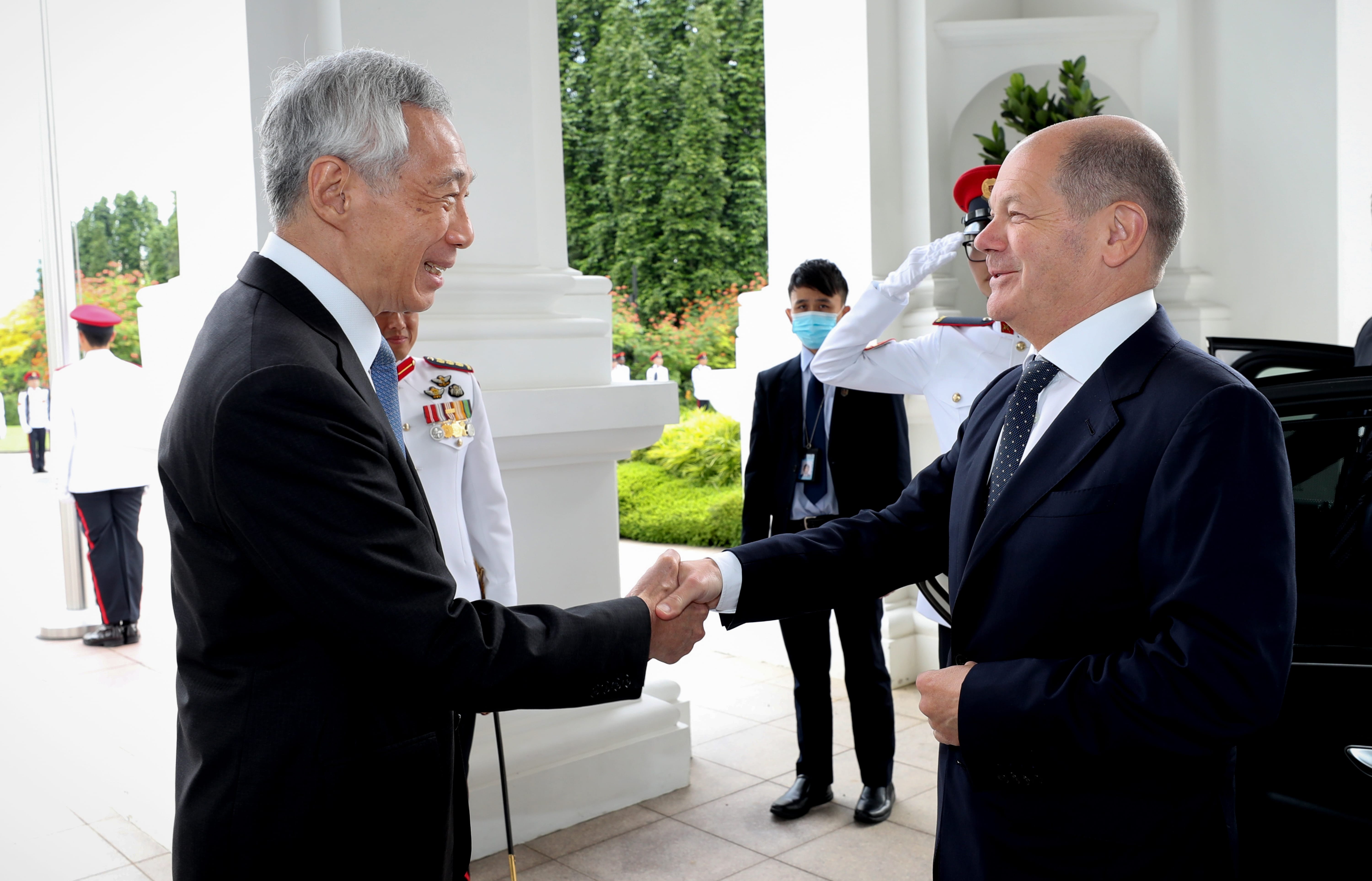 Lee Hsien Loong and Olaf Scholz shaking hands, guards nearby, at a white building.