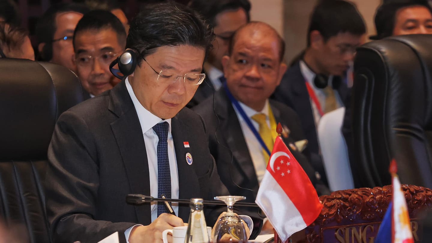 Man in suit with headset writes at table with Singapore flag and others in background.