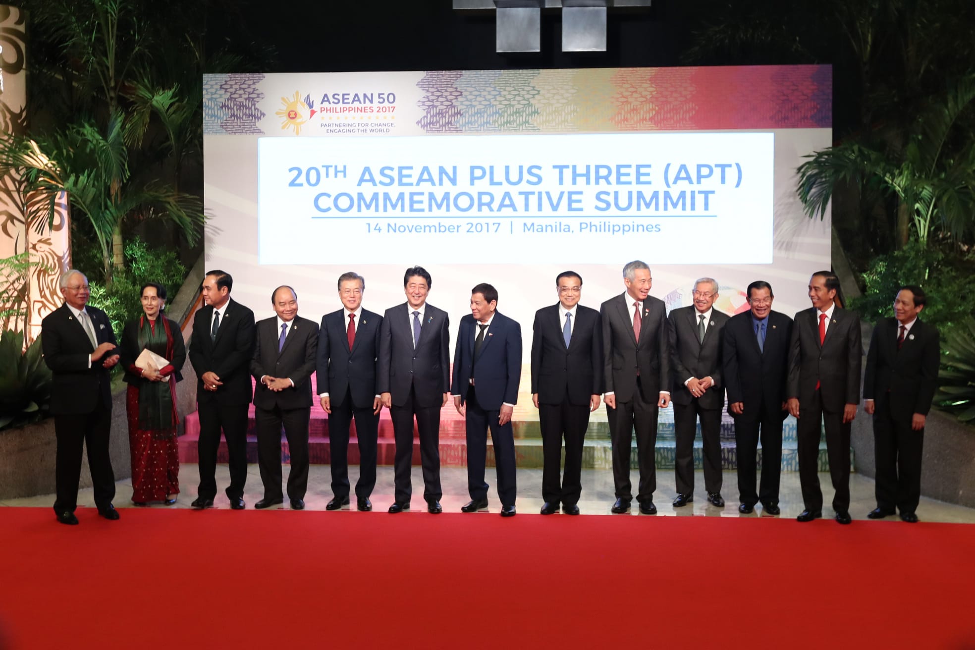 World leaders in suits stand on a red carpet in front of the "20th ASEAN Plus Three" summit backdrop.