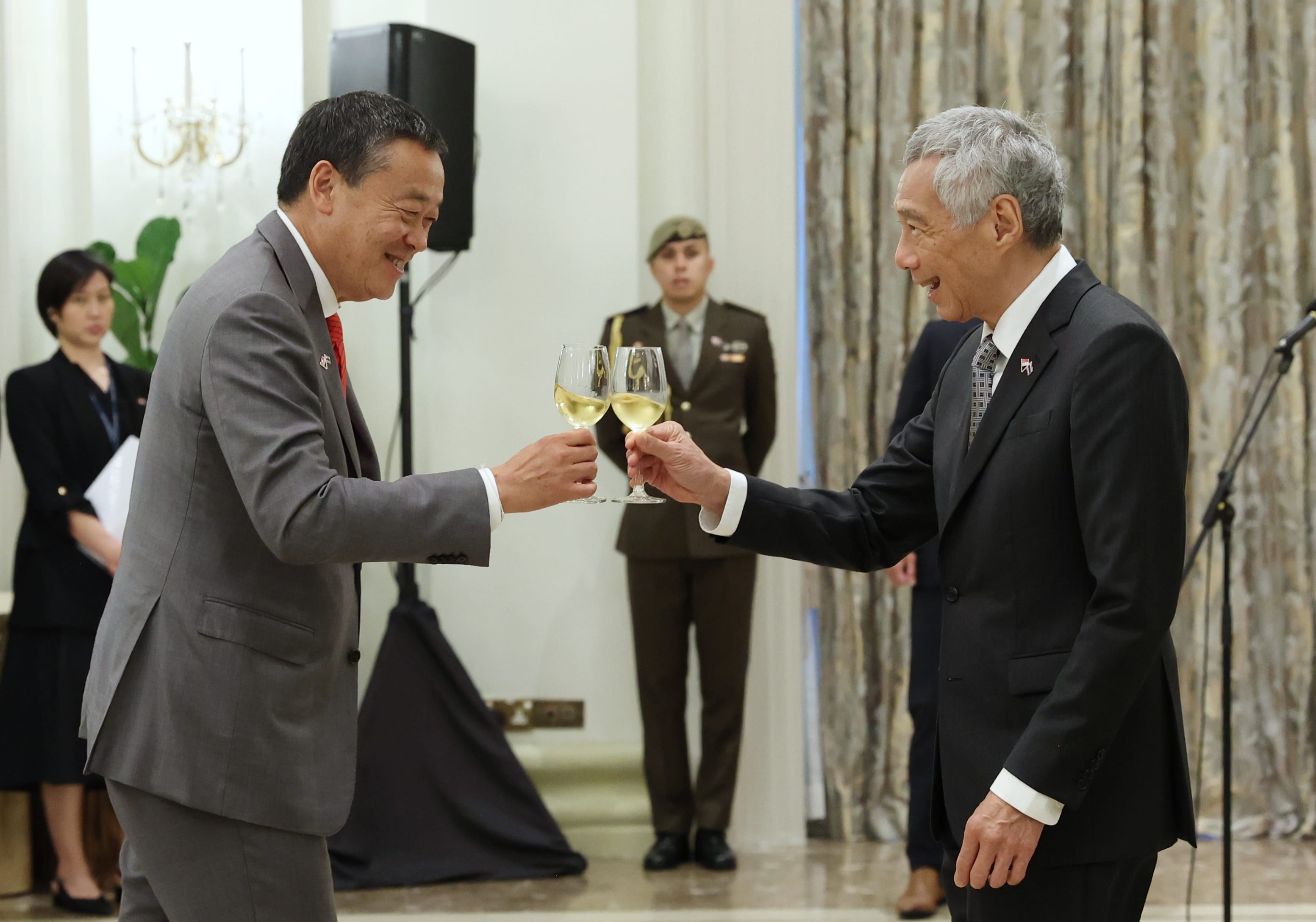 Two men in suits toasting with wine glasses, uniformed person in background.