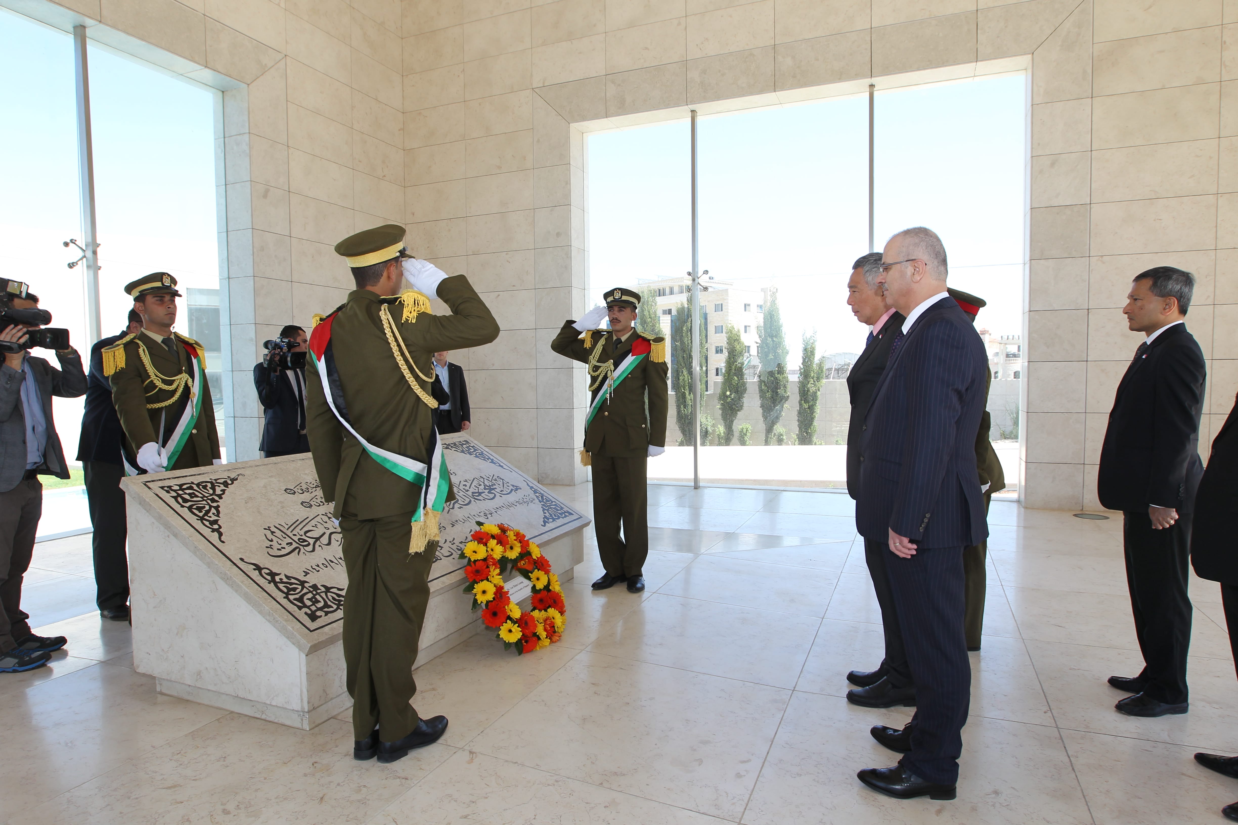 Soldiers in green uniforms salute a tomb with Arabic writing; men in suits stand nearby.