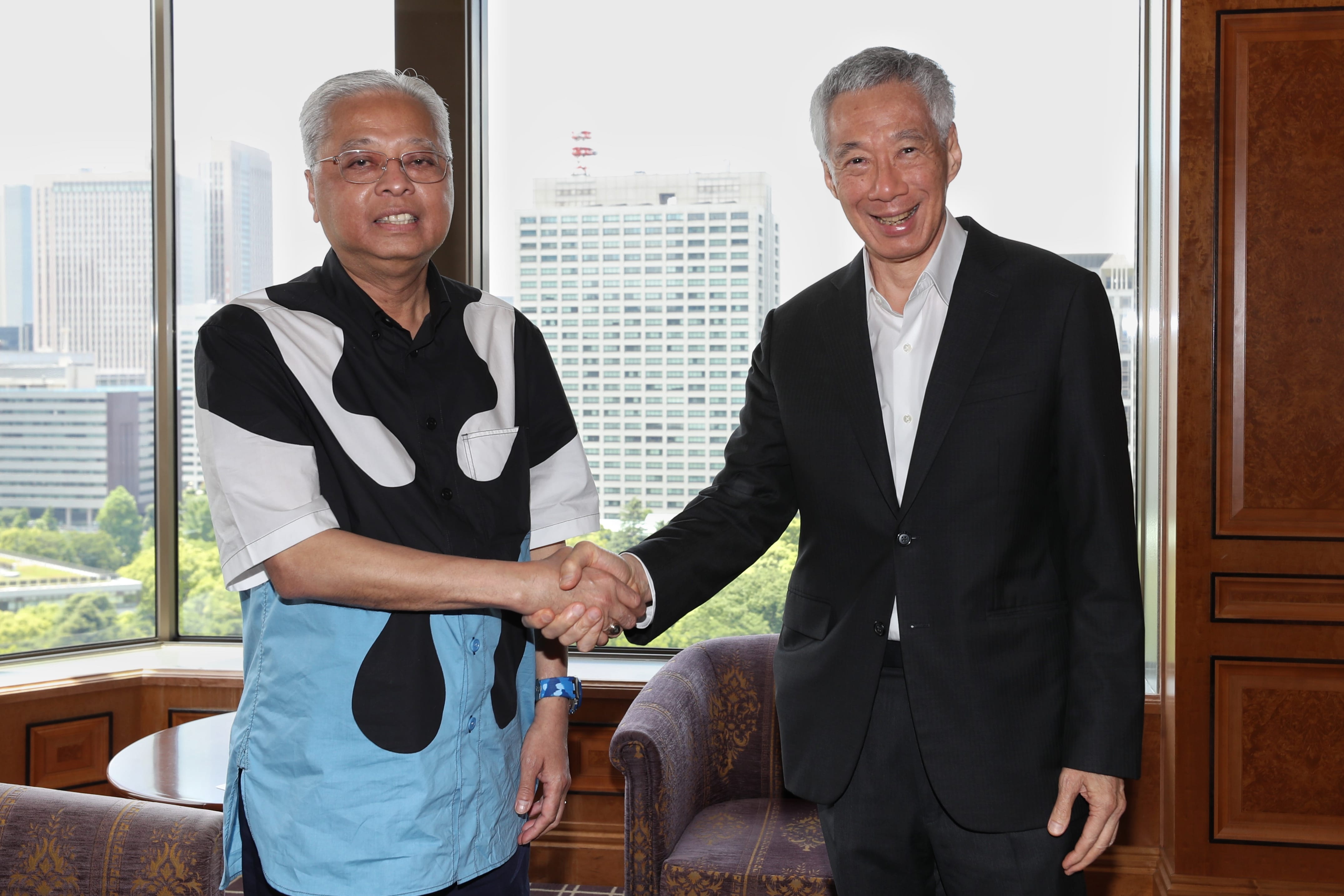 Lee Hsien Loong shaking hands with man in patterned shirt, cityscape visible outside window.