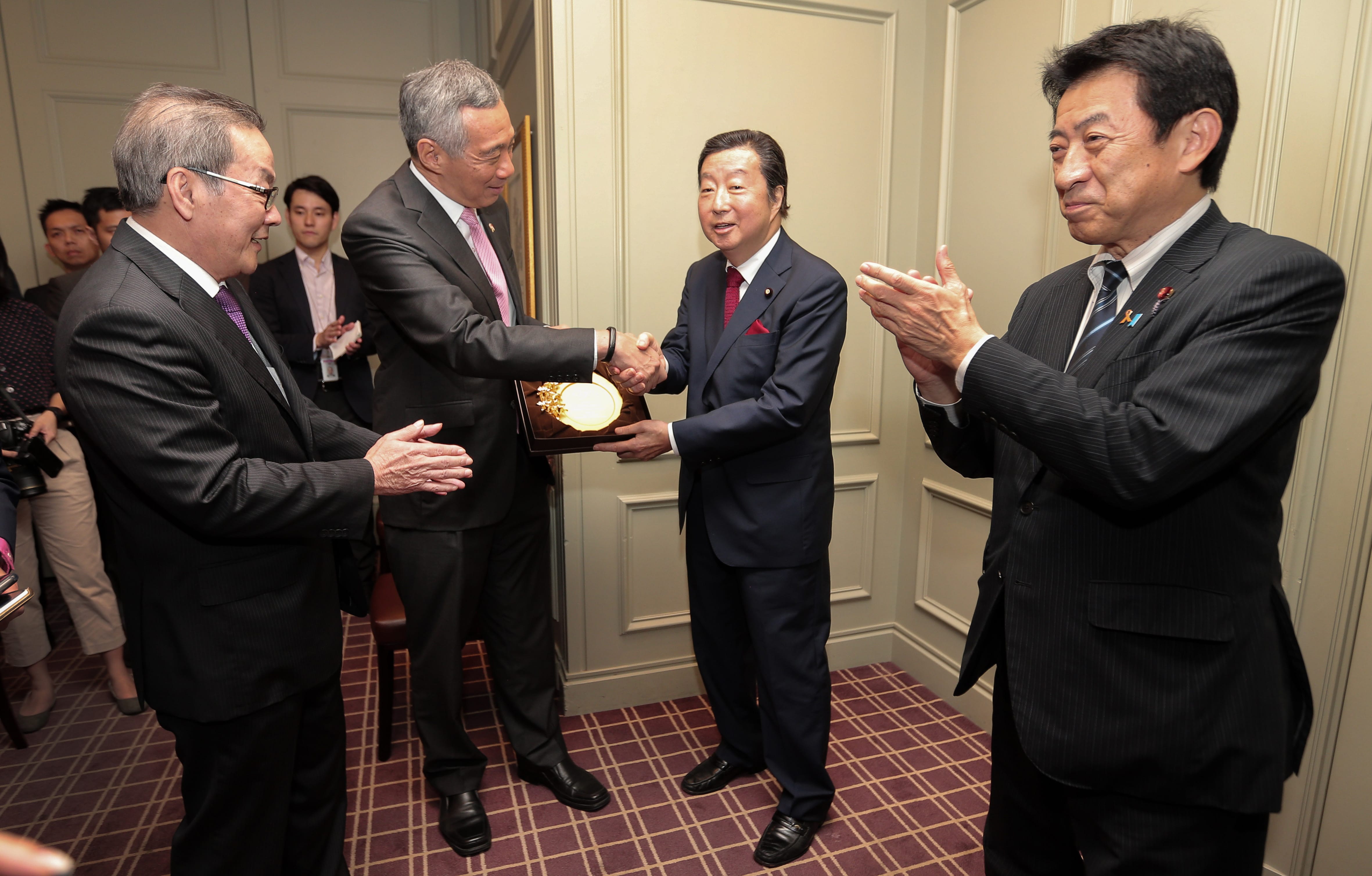 Lee Hsien Loong shaking hands, receiving a golden plaque; other men in suits observe and applaud.