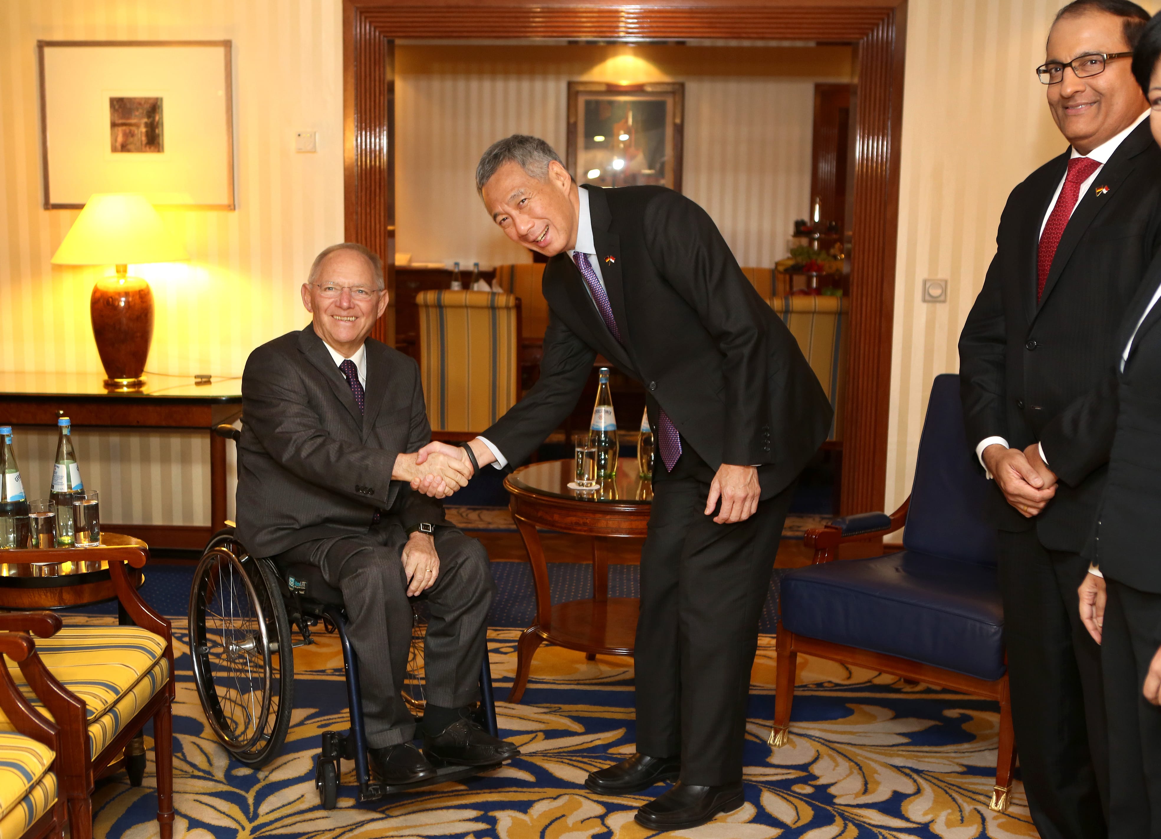 Lee Hsien Loong shakes hands with Wolfgang Schäuble in a wheelchair. Two men stand nearby.