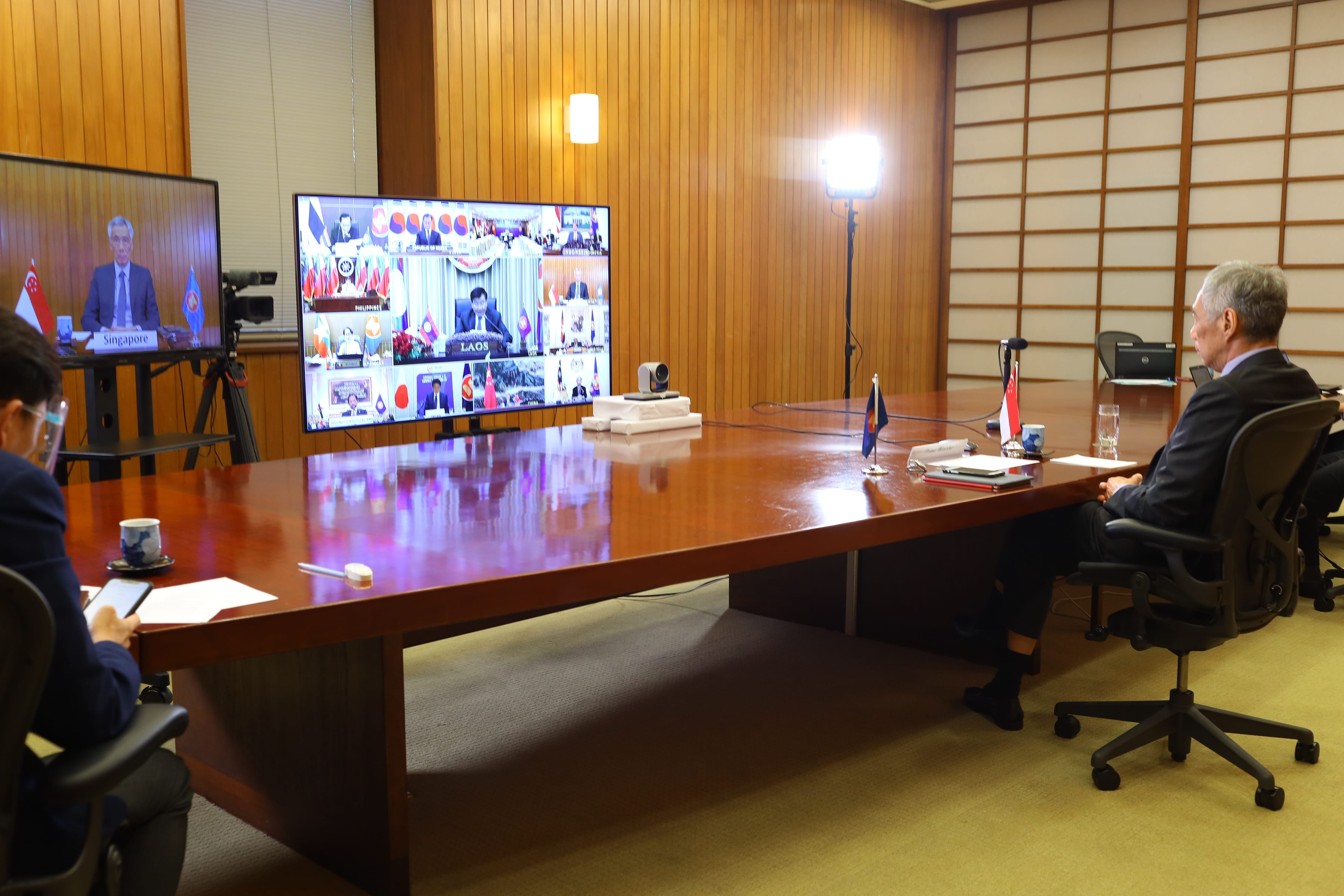 Man at a large table, video conferencing on monitors, Singapore flag visible.