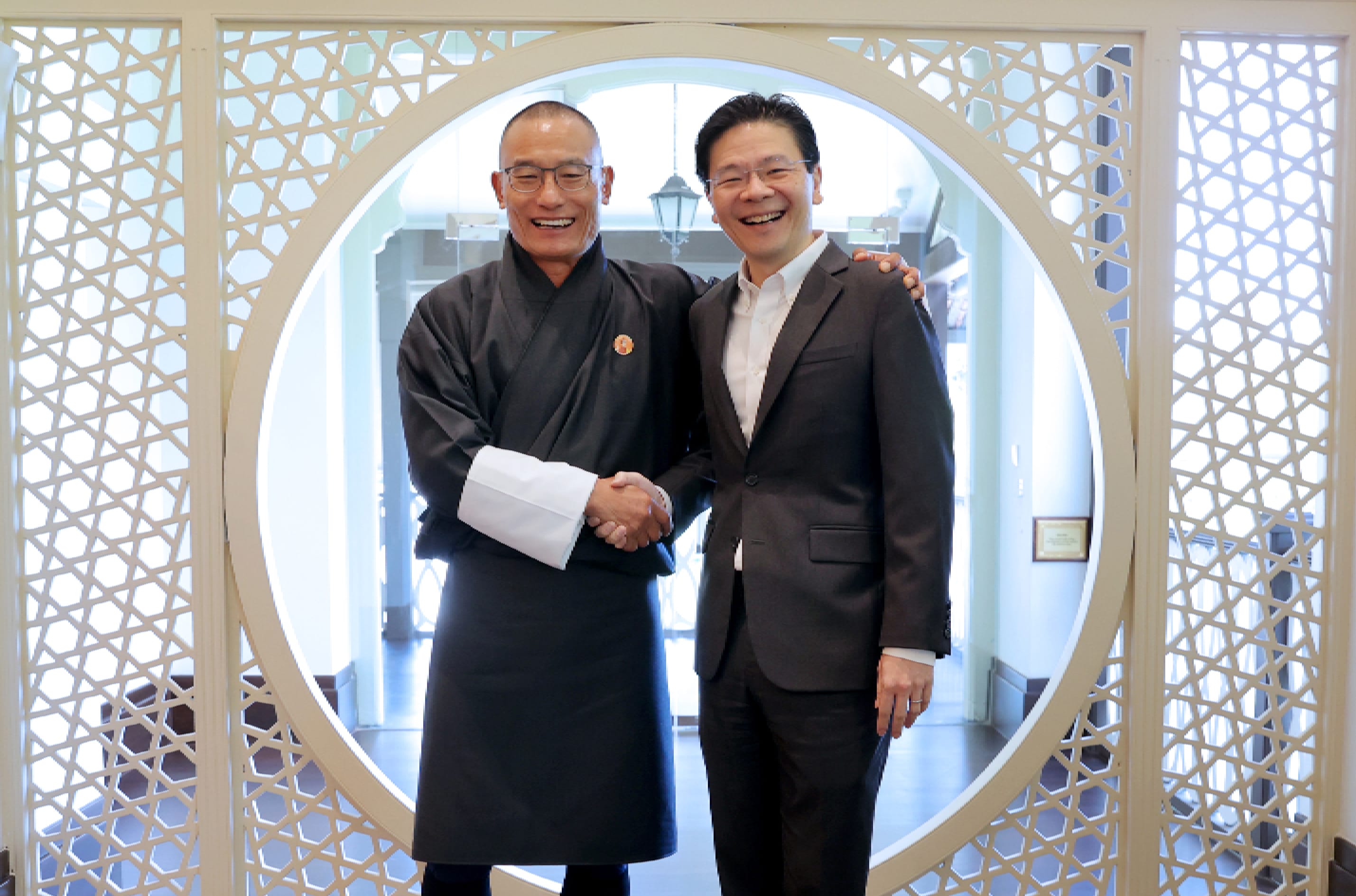 Two men shake hands in front of a round window with a geometric pattern. One wears a suit, the other a dark traditional robe.