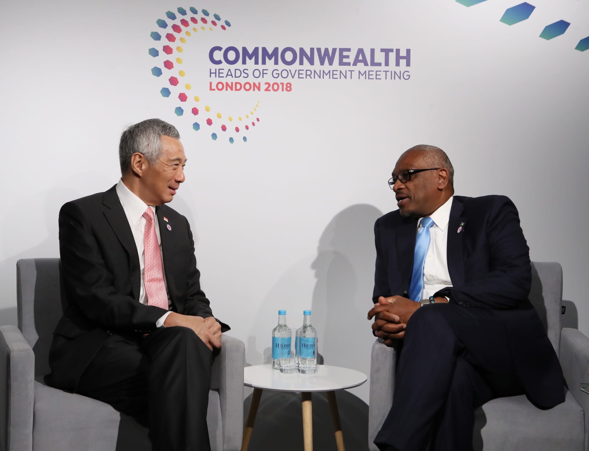 Two men in suits sit facing each other against a "Commonwealth" backdrop. "Hildon" water bottles on a table.