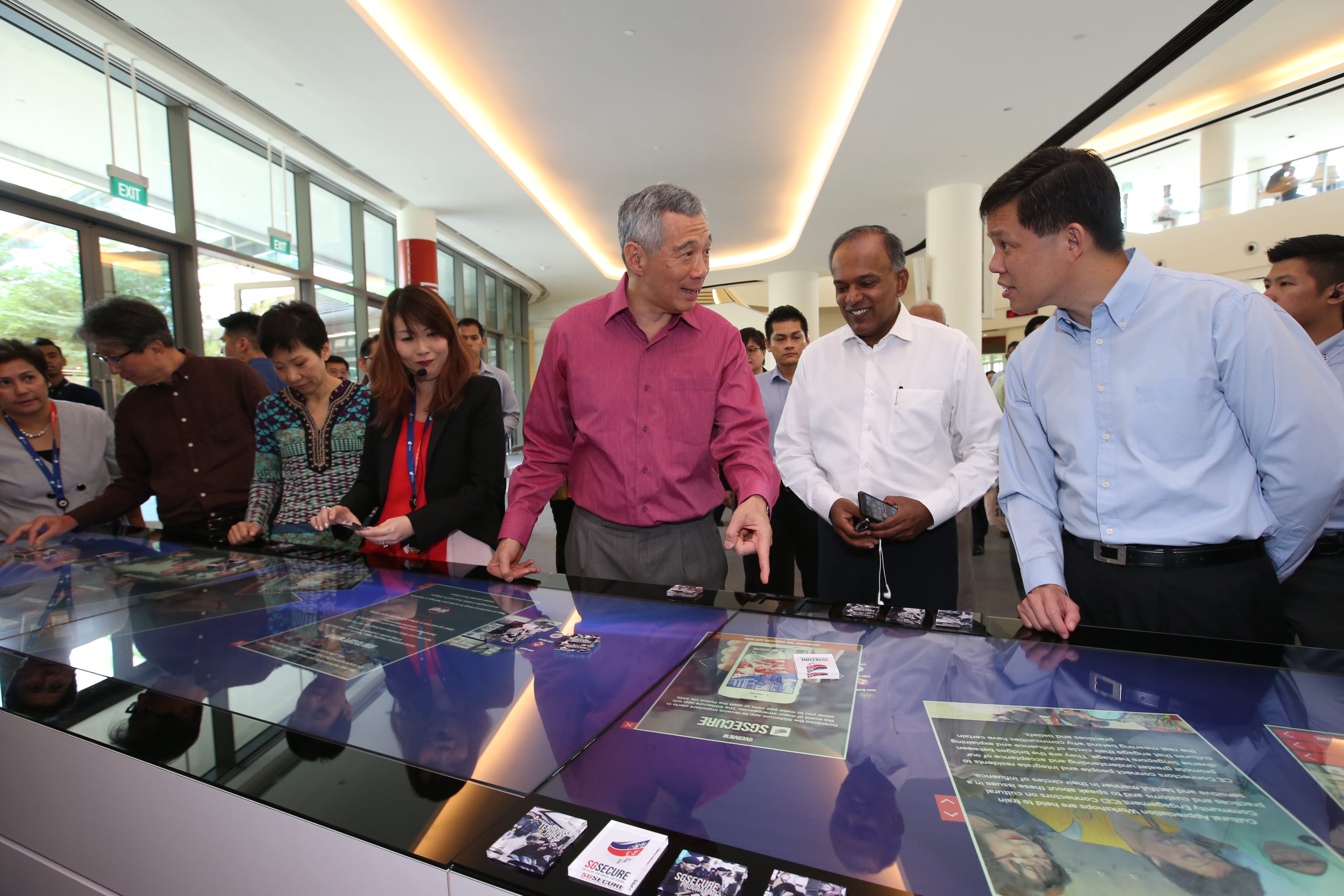 Lee Hsien Loong and others examine interactive digital displays with SG Secure branding in a brightly lit interior.