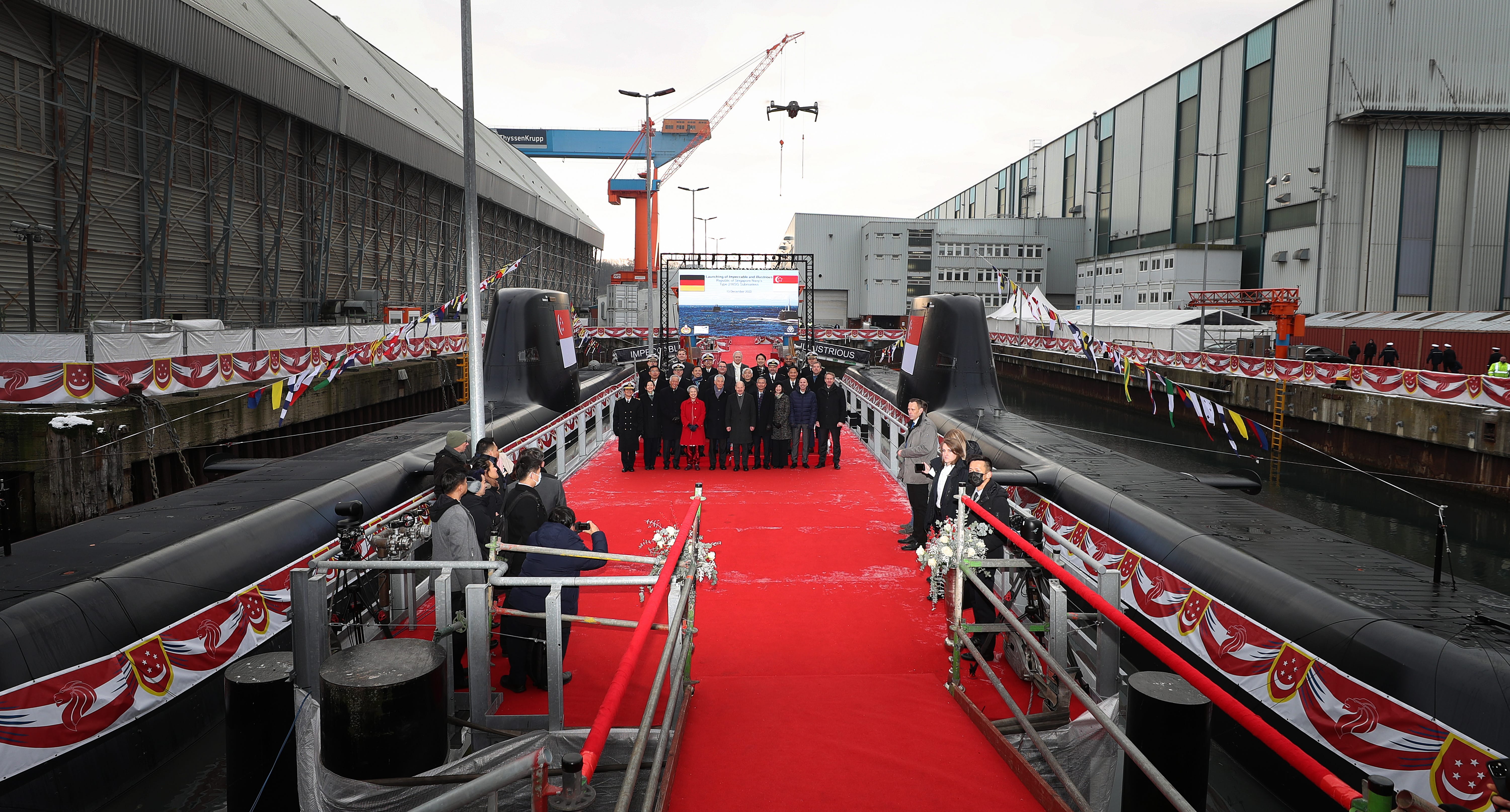 Ceremony on submarines with red carpet, flags, and groups of people in a shipyard.