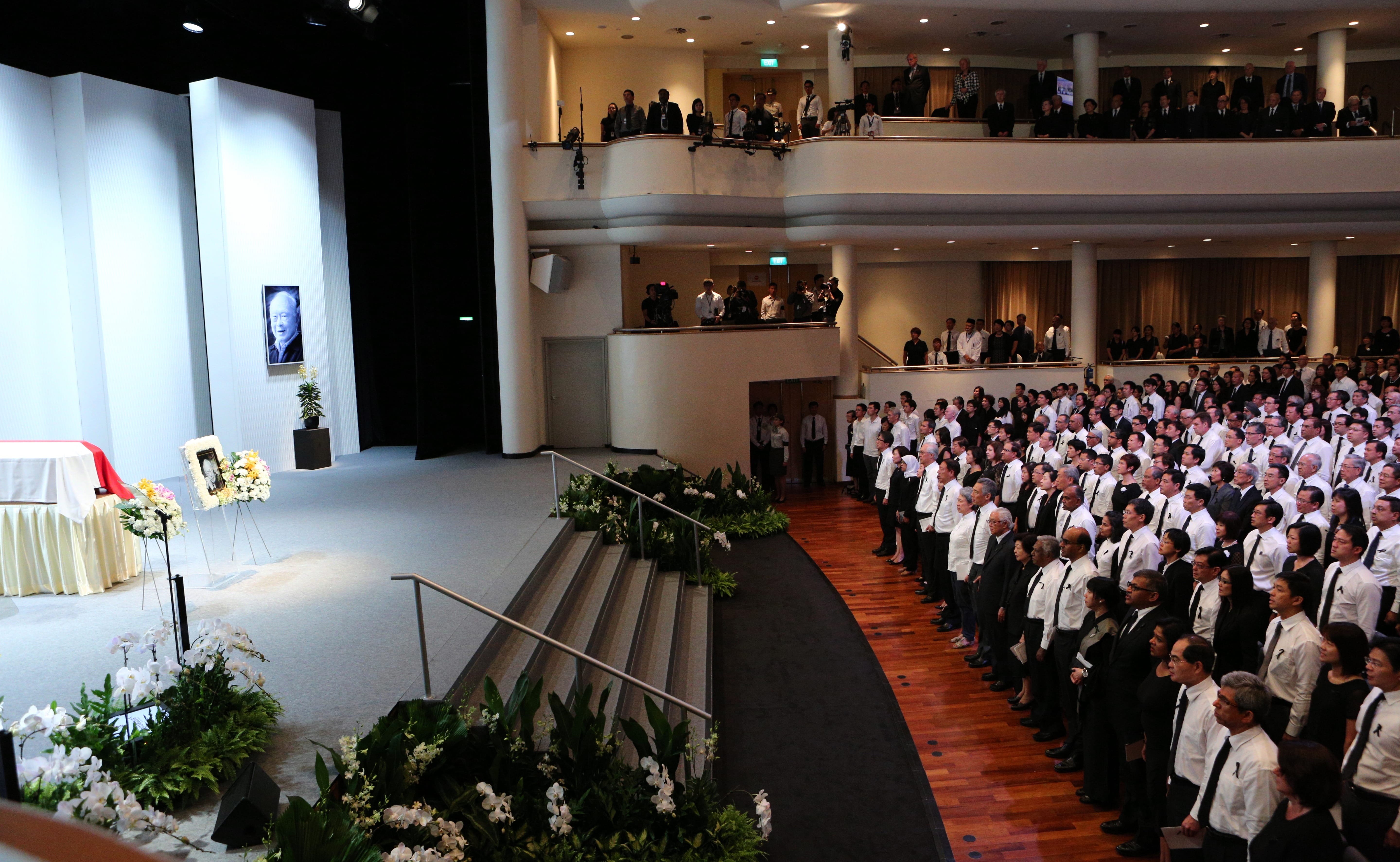 Auditorium with attendees, casket on stage, framed portrait, and floral arrangements. Balconies line the upper levels.