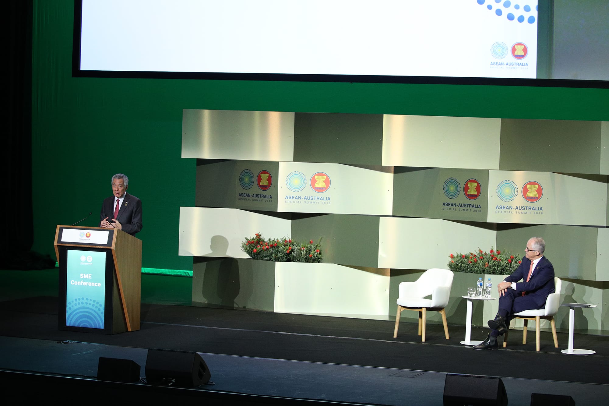 Two men on stage at ASEAN-Australia SME Conference, one speaking at a podium.