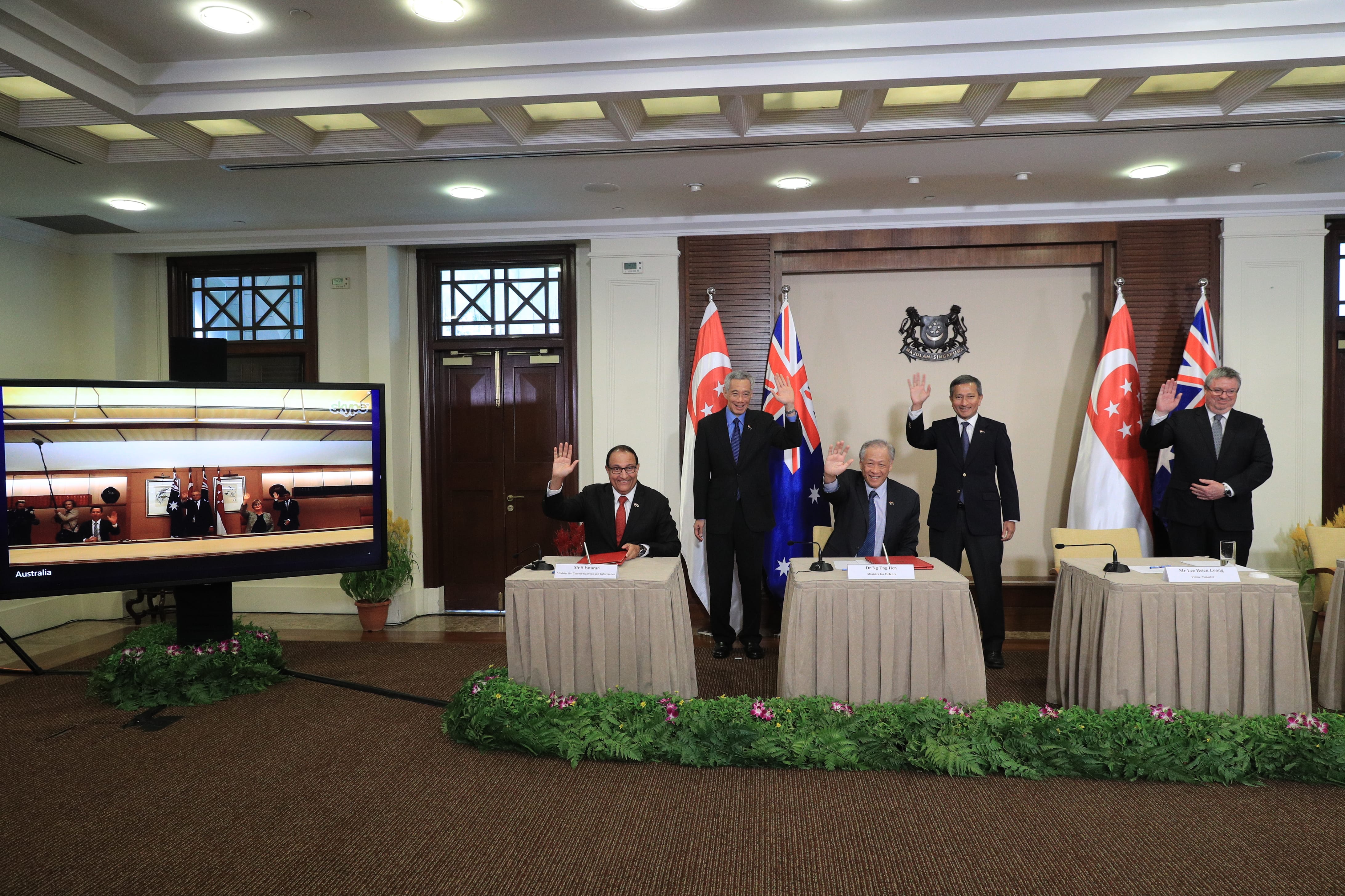 Four men in suits waving, standing behind draped tables with flags and a Singapore coat of arms. A video screen shows similar scene.
