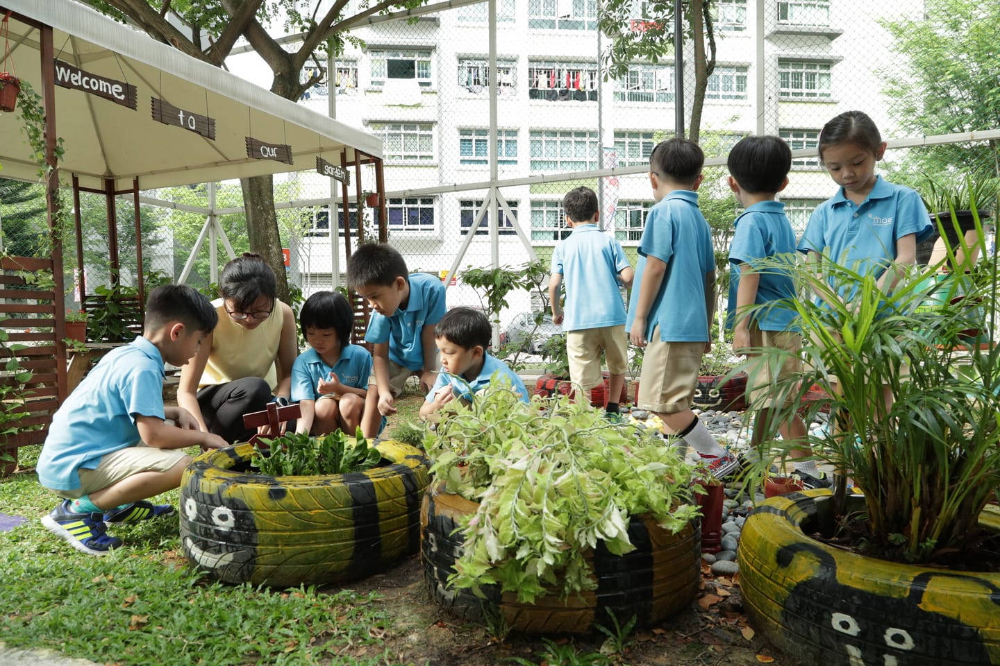 Kids in blue shirts, plus teacher, tend plants in painted tire planters outside a building.