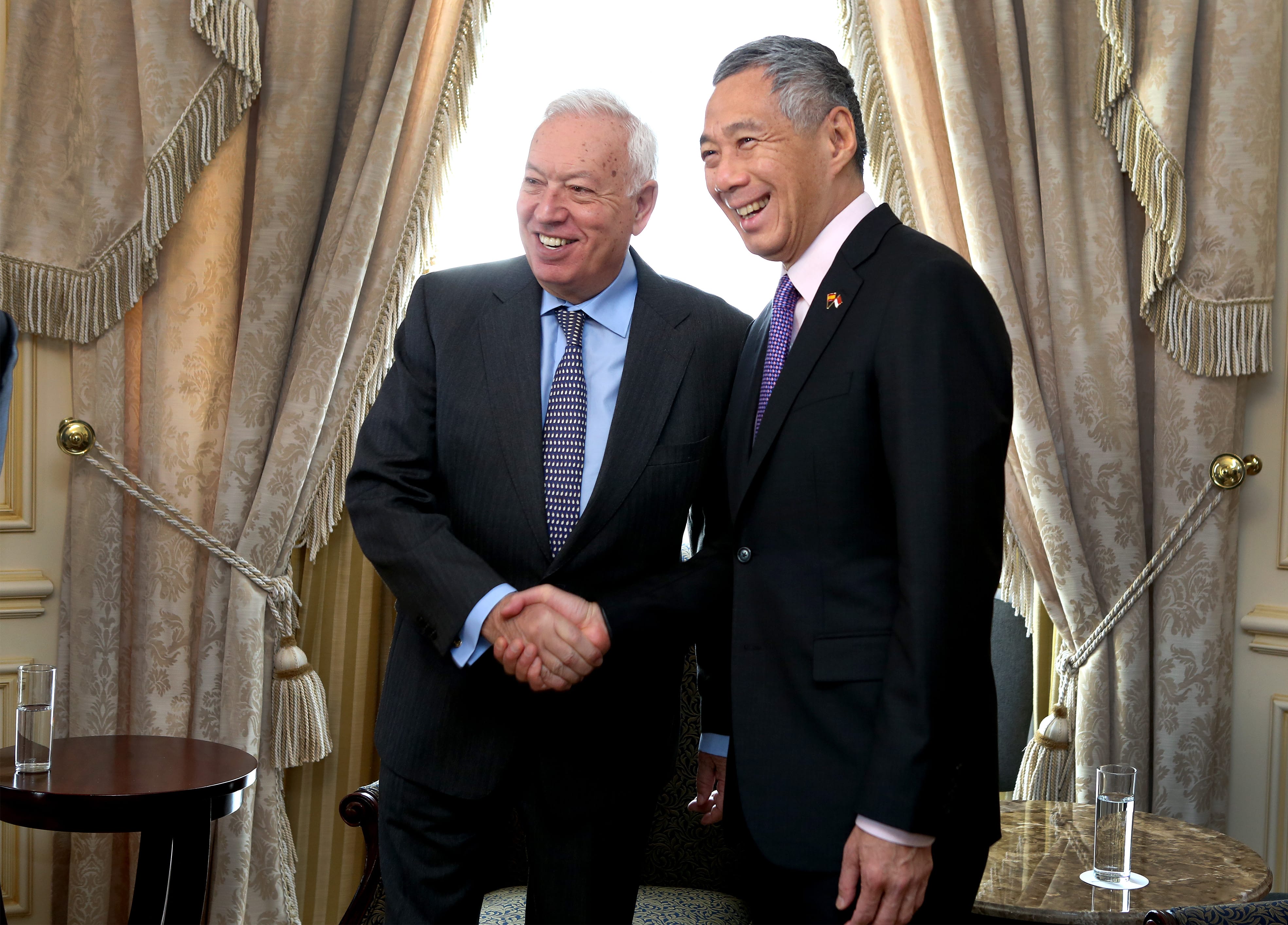 Two men in suits, shaking hands and smiling, stand before patterned curtains. Lee Hsien Loong is on the right.
