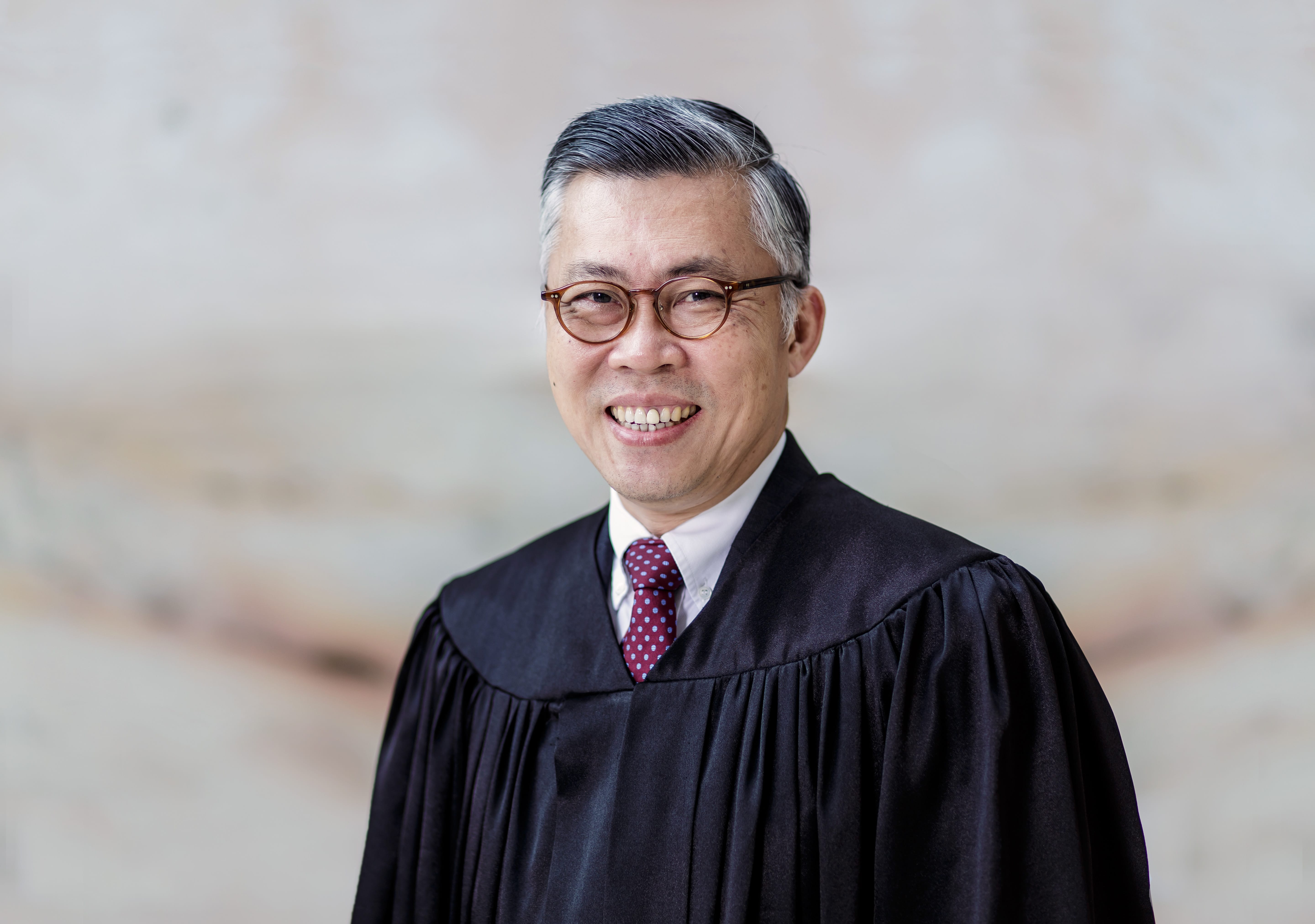 Person in a black judicial robe and glasses, smiling. Red tie with white dots.