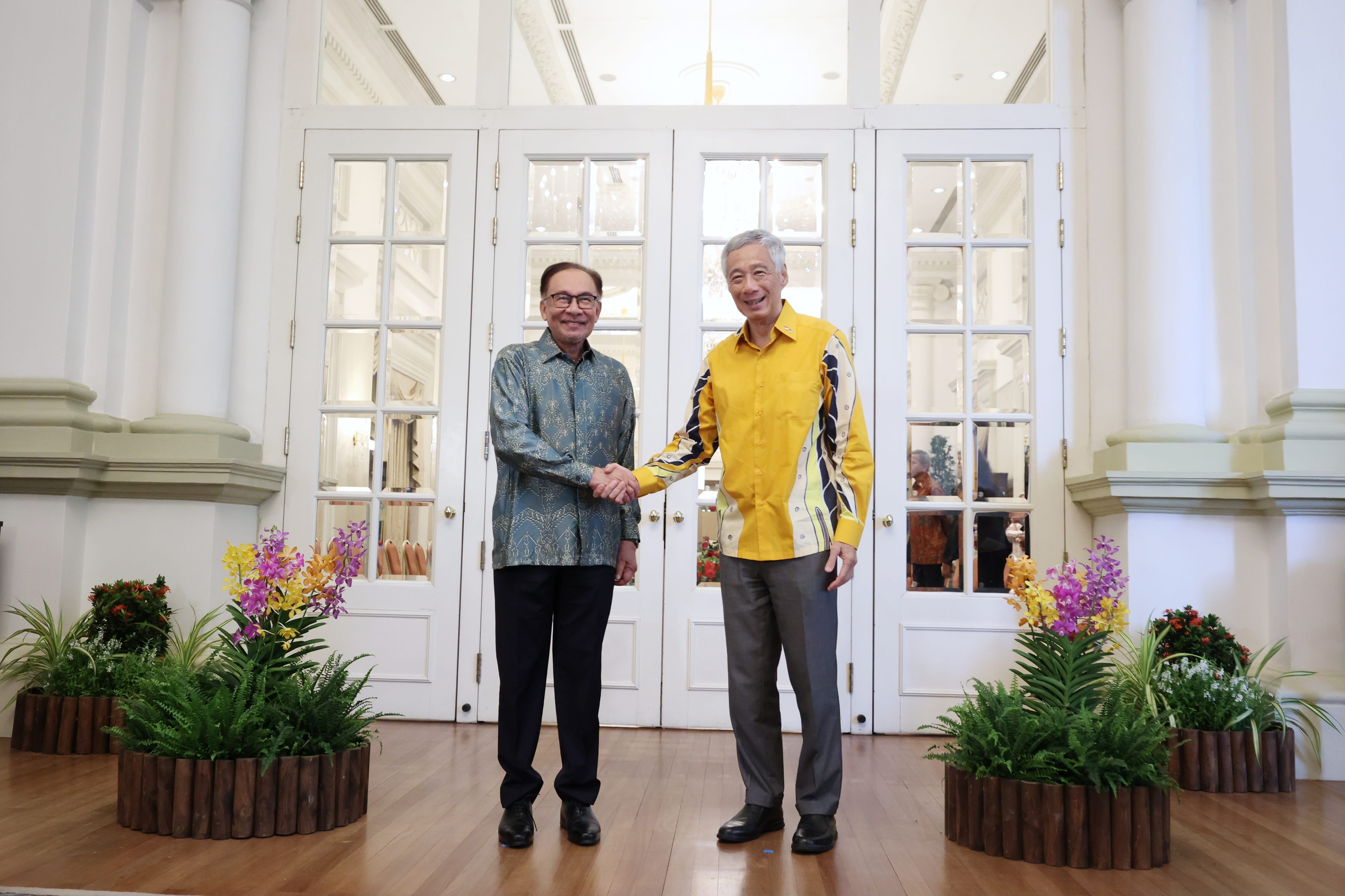 Anwar Ibrahim and Lee Hsien Loong shake hands in front of glass doors, beside floral planters.