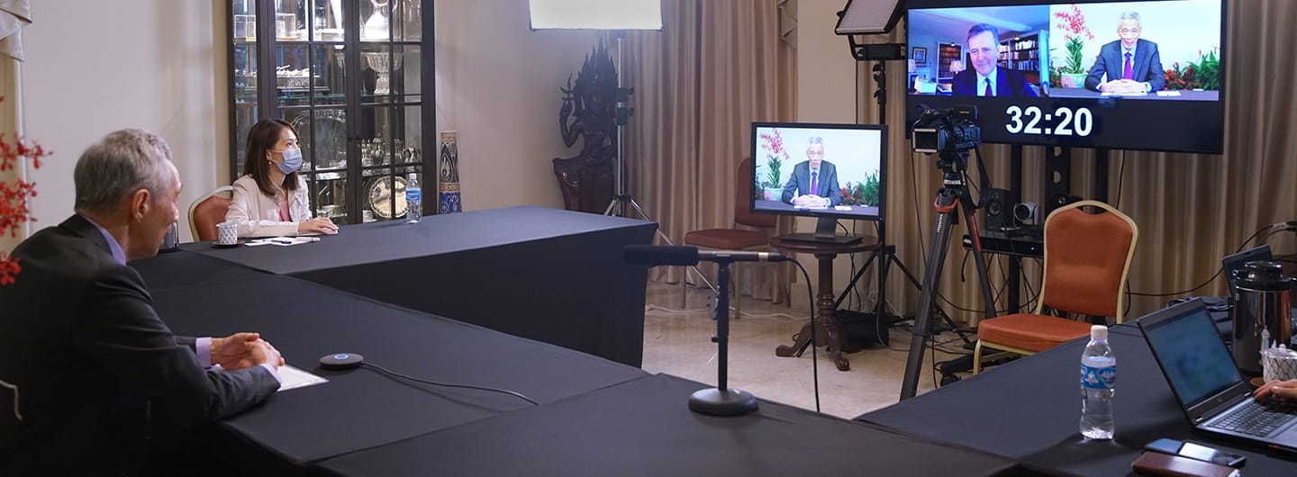 In-person/remote meeting setup: two people, screens showing remote participants, mics, cameras.