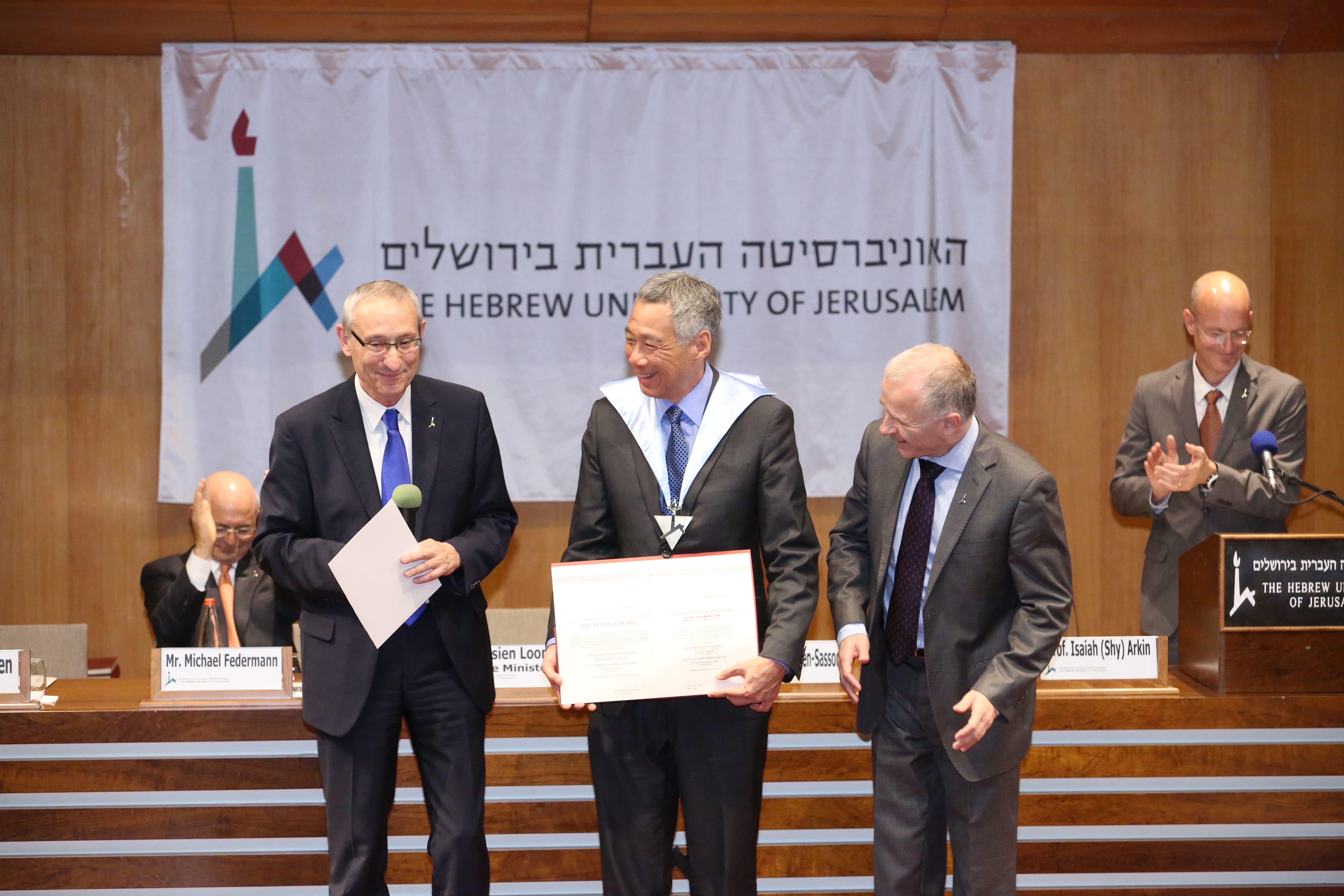 Three men onstage in suits; central figure in academic gown holding award from Hebrew University of Jerusalem.