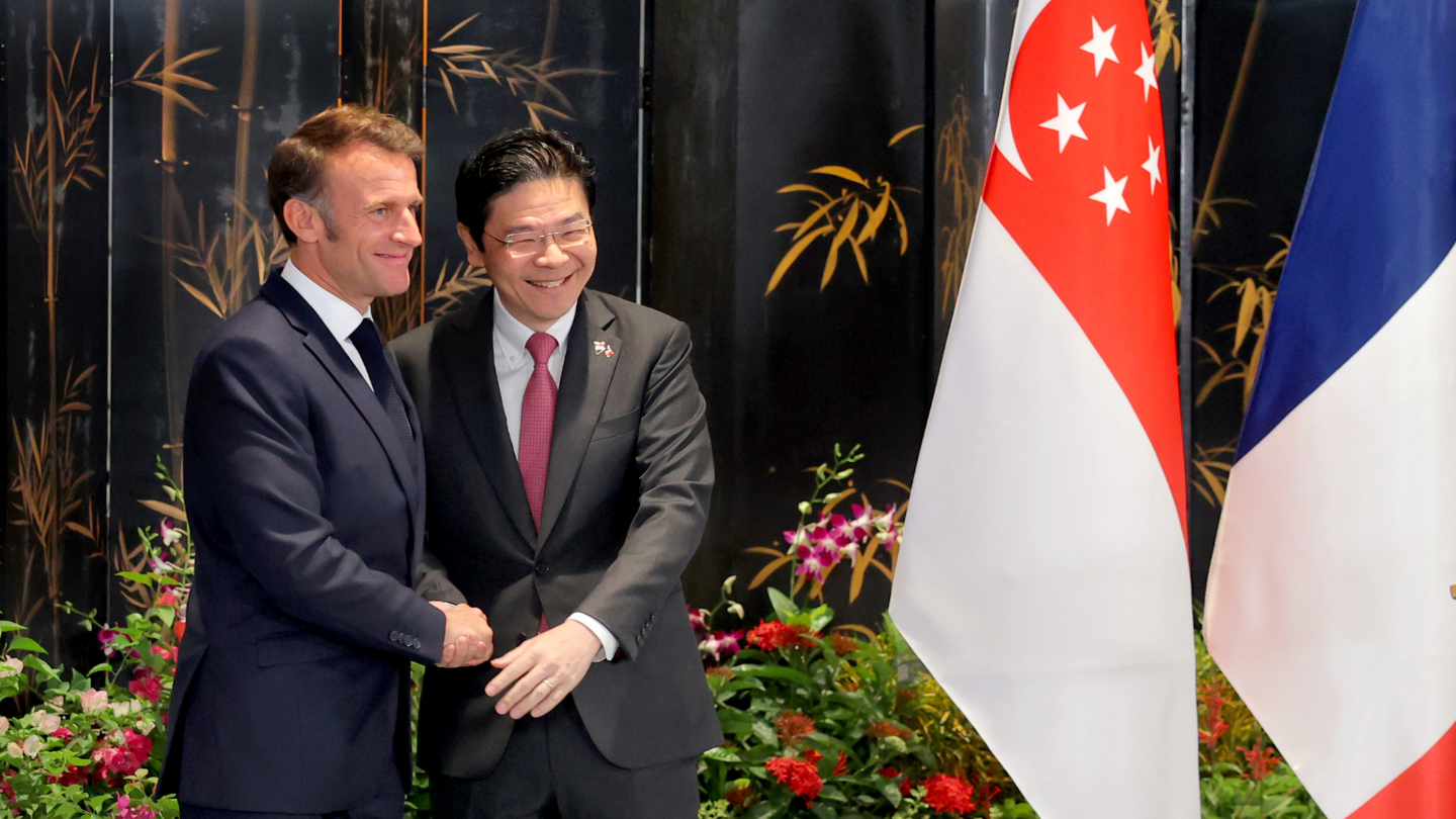 Emmanuel Macron shaking hands with a man in front of Singapore and France flags.