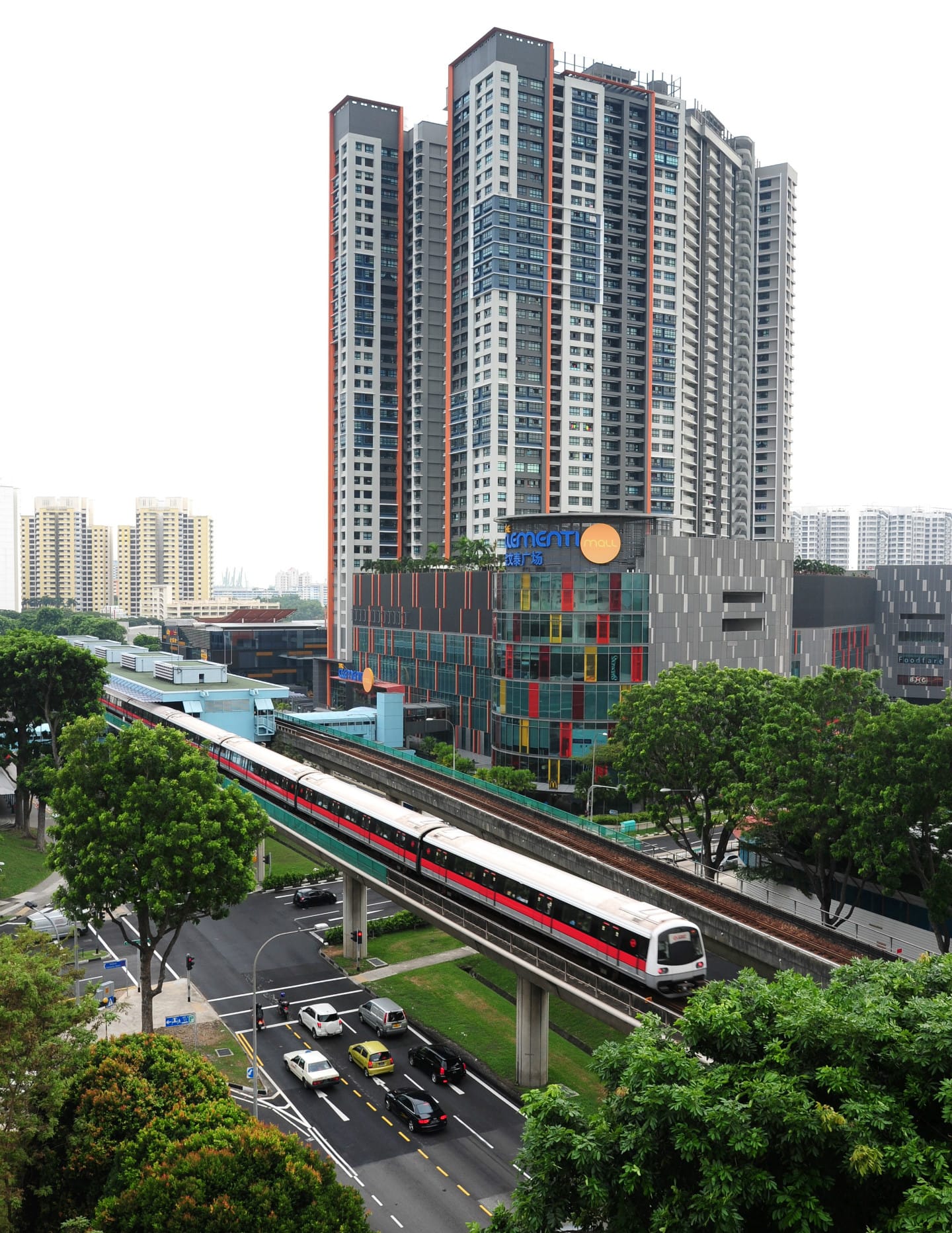 Elevated train with red stripe moving past buildings, trees, and a road with traffic.