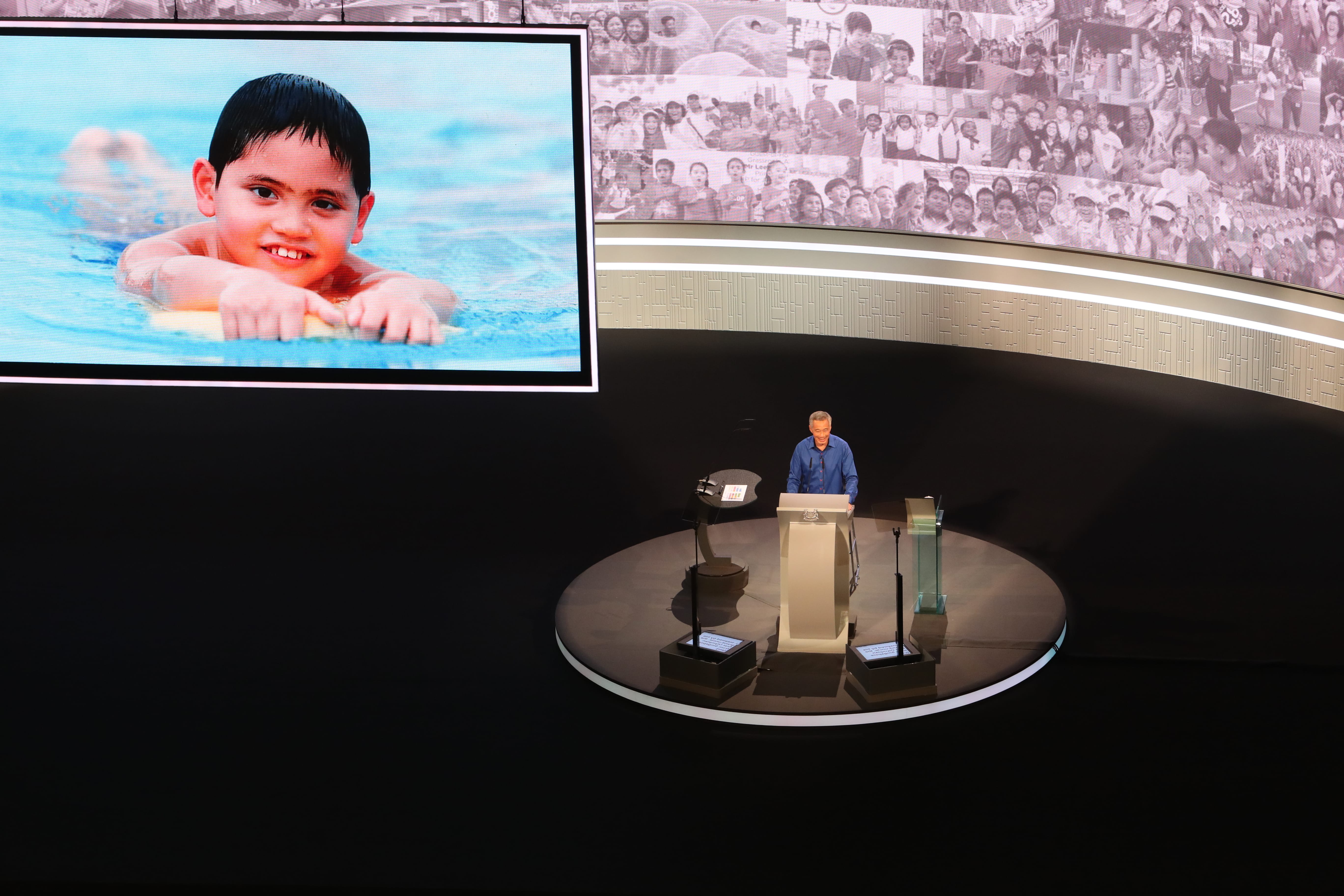 Lee Hsien Loong at podium, large screen shows child swimming; crowd background.