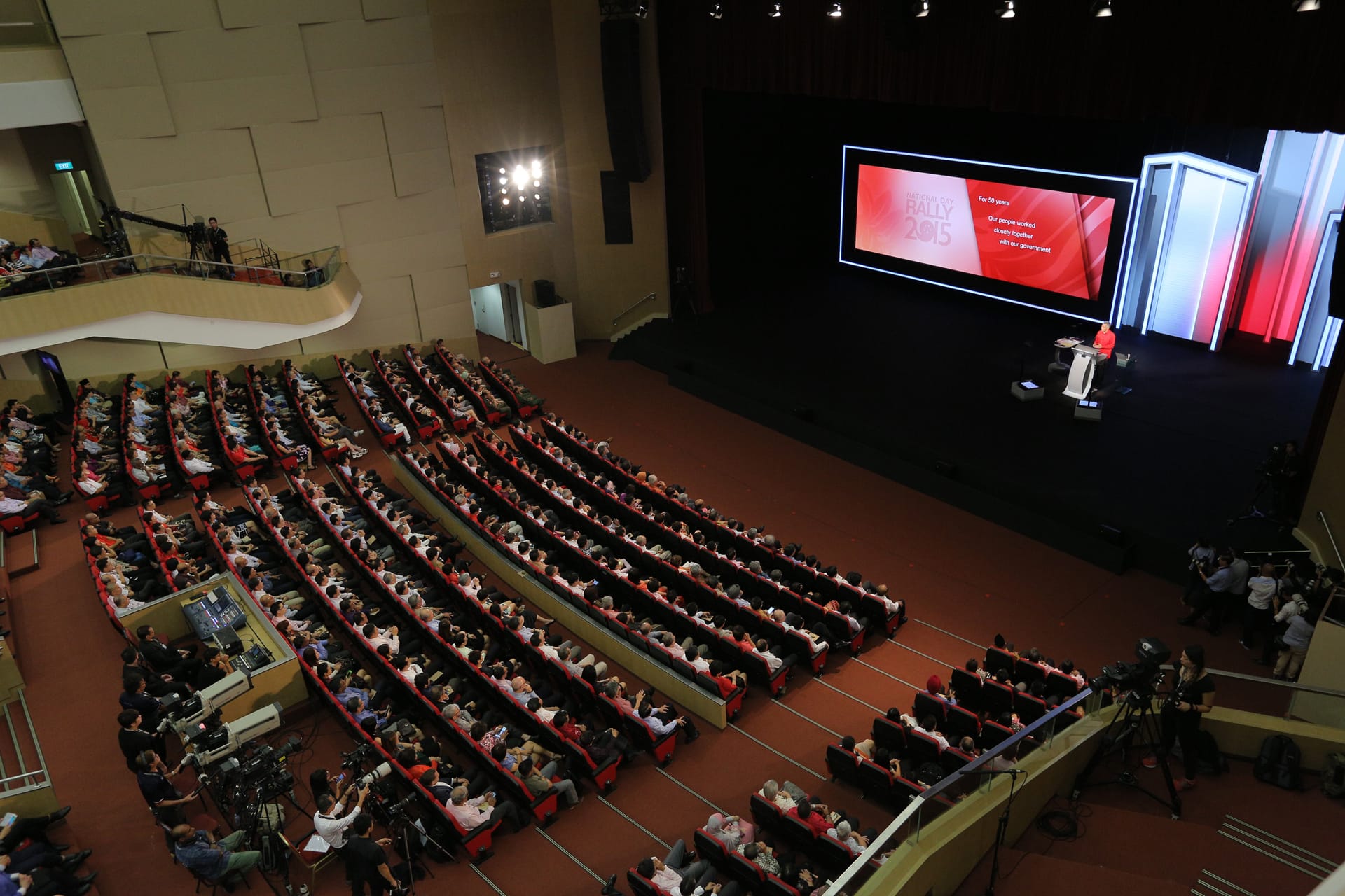 Auditorium filled with people facing stage, screen reads "National Day Rally 2015".
