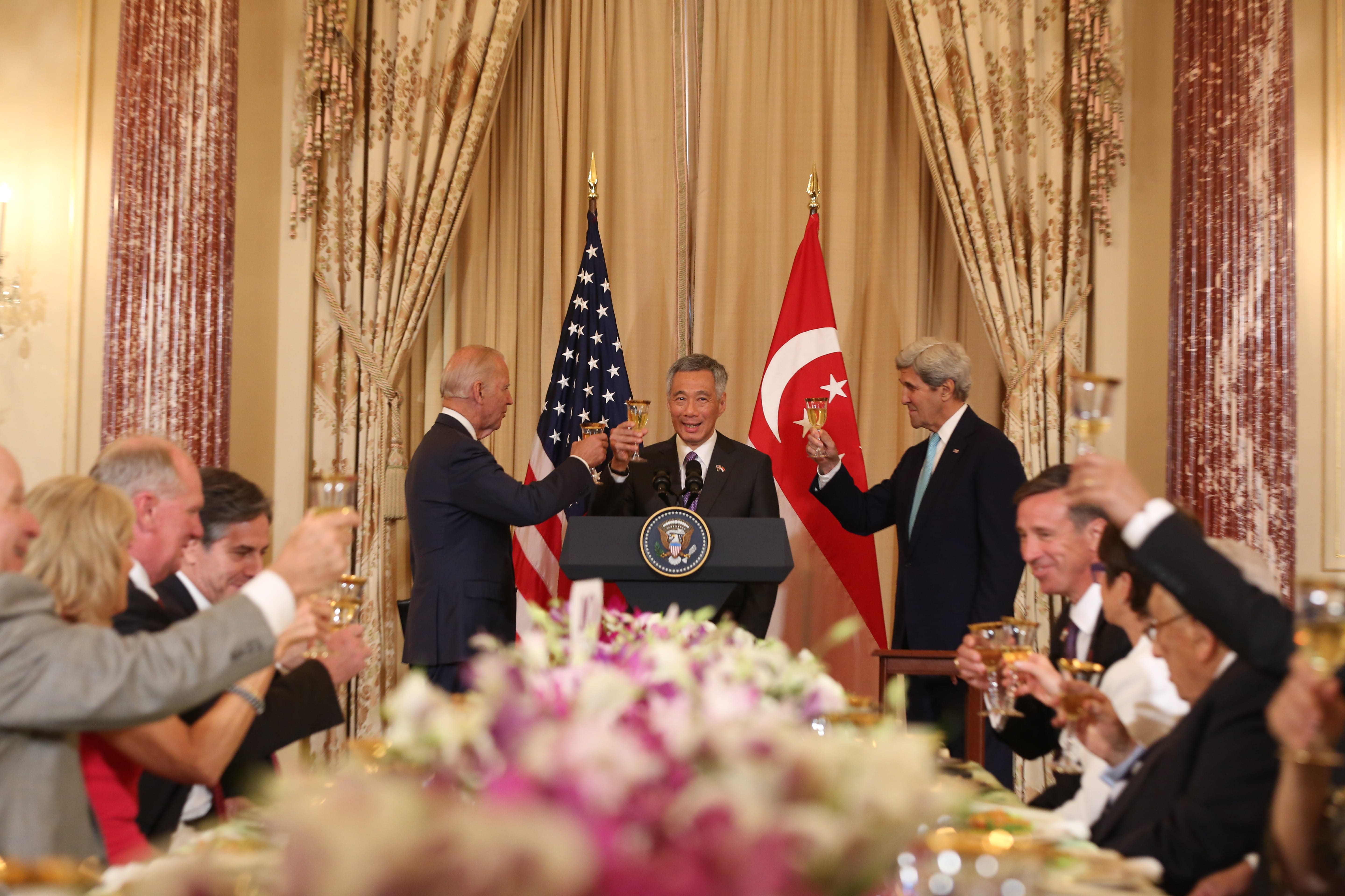 Joe Biden, Lee Hsien Loong, and John Kerry toasting at a formal dinner with flags behind them.