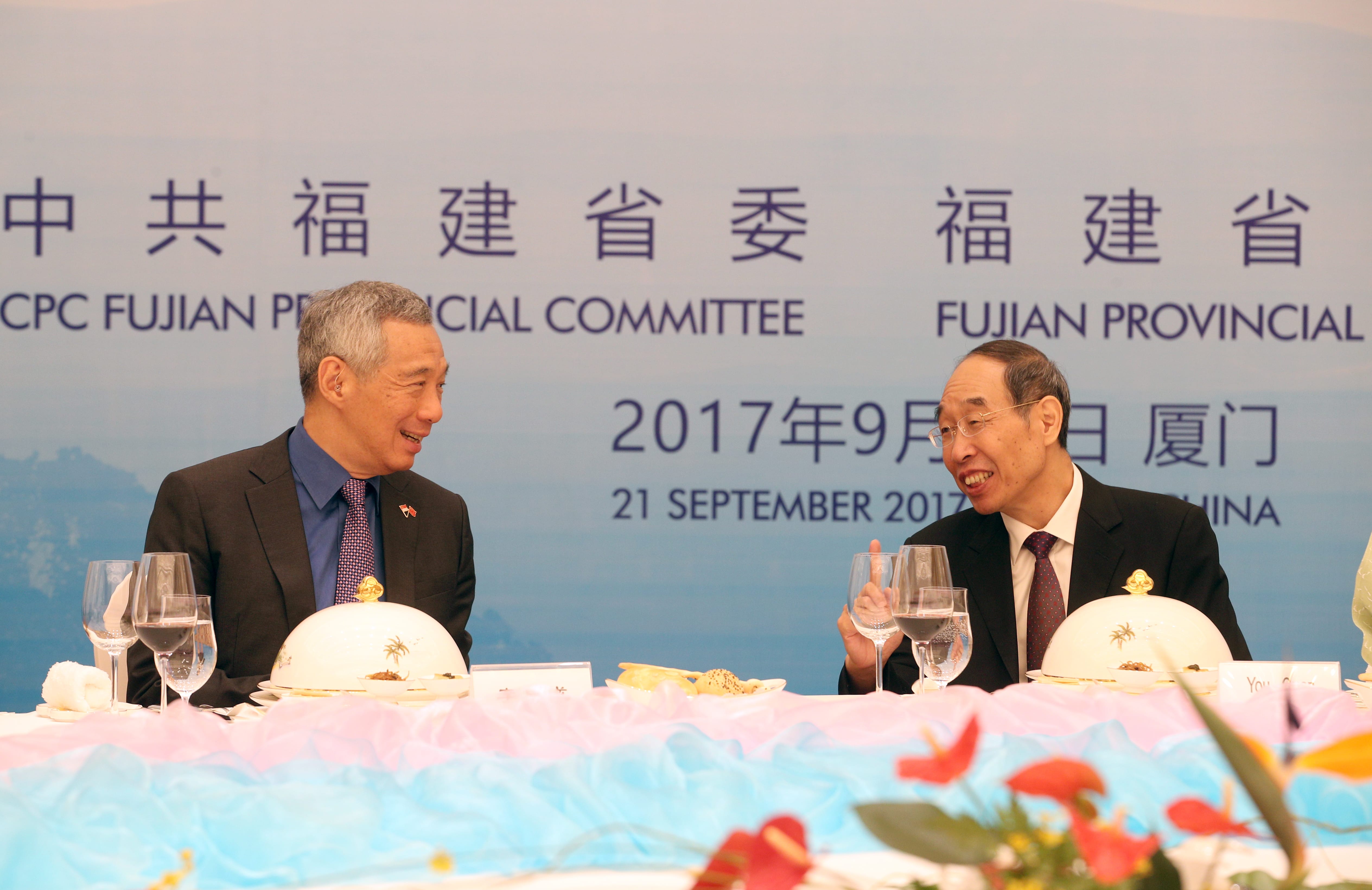 Lee Hsien Loong and another man at a table with wine glasses and text backdrop.