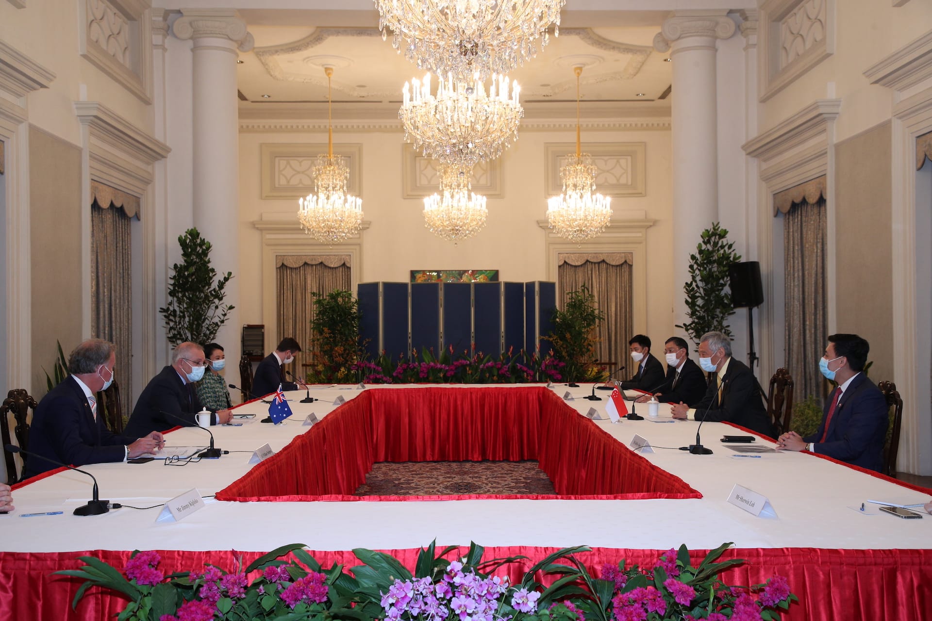 People at long table with flags, wearing masks, under chandeliers in ornate room.