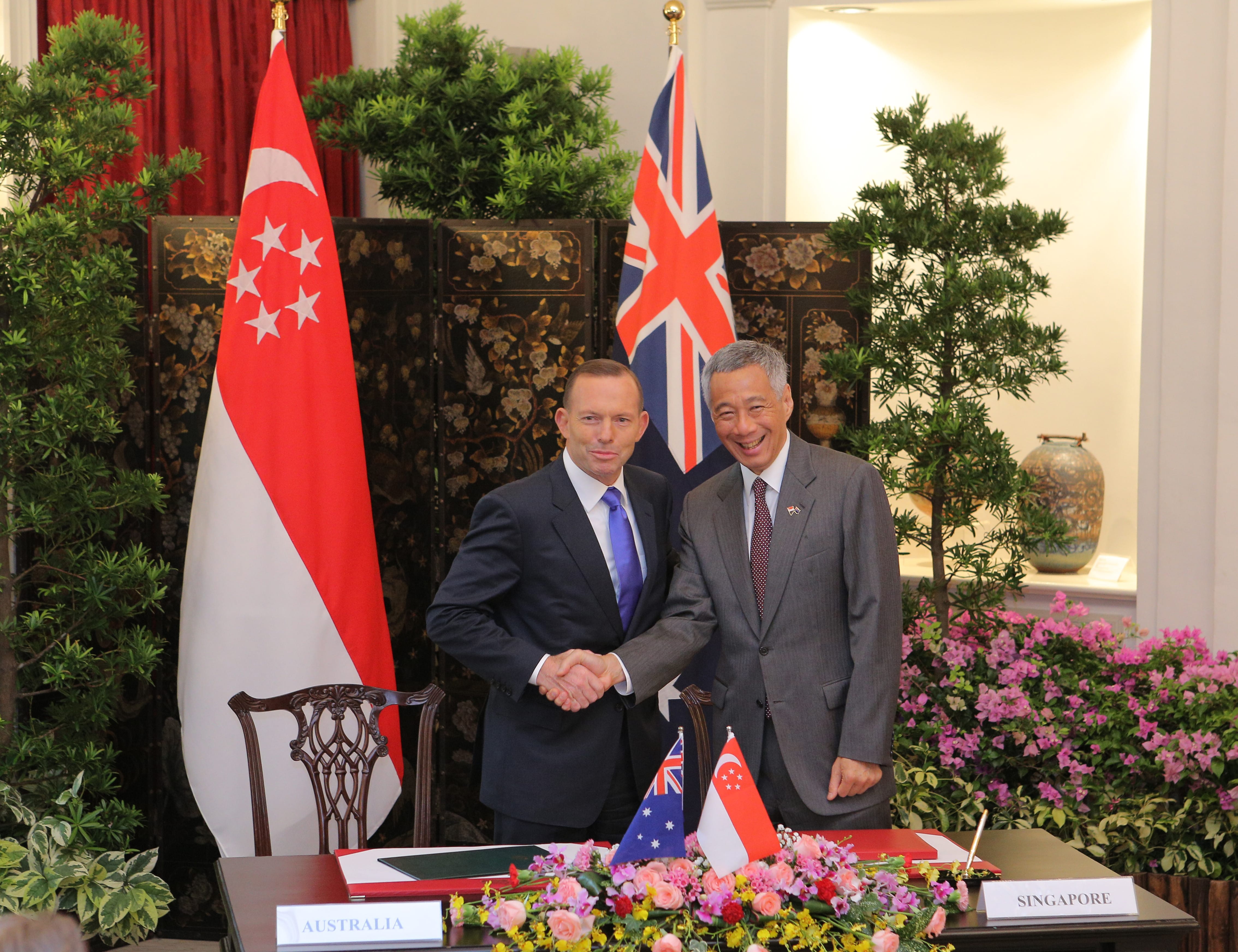 Two men in suits shake hands near flags of Singapore, UK, and Australia, plus floral arrangements.