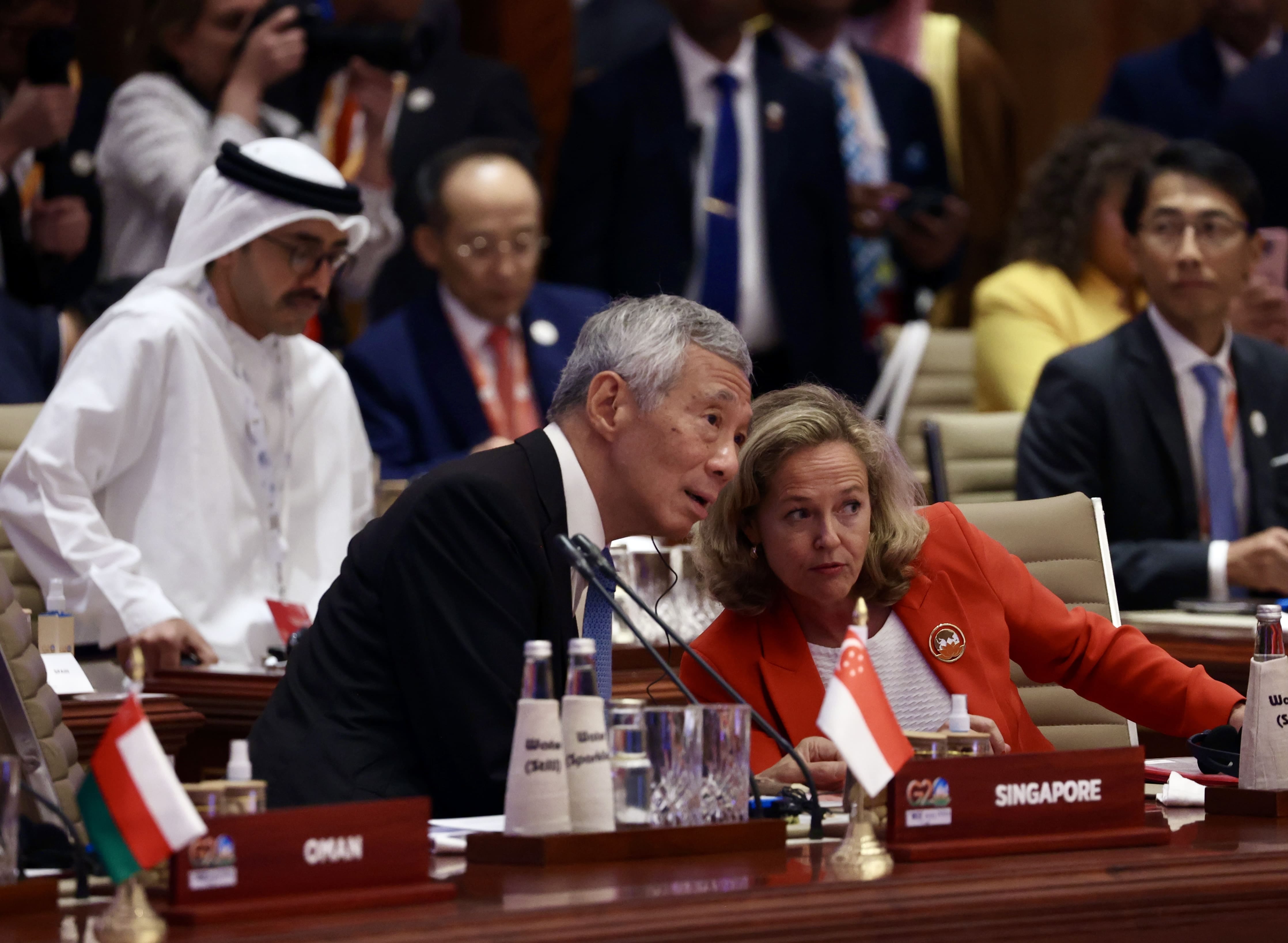 Lee Hsien Loong and a woman in an orange blazer at a G20 meeting. The Singapore flag and nameplate are visible.