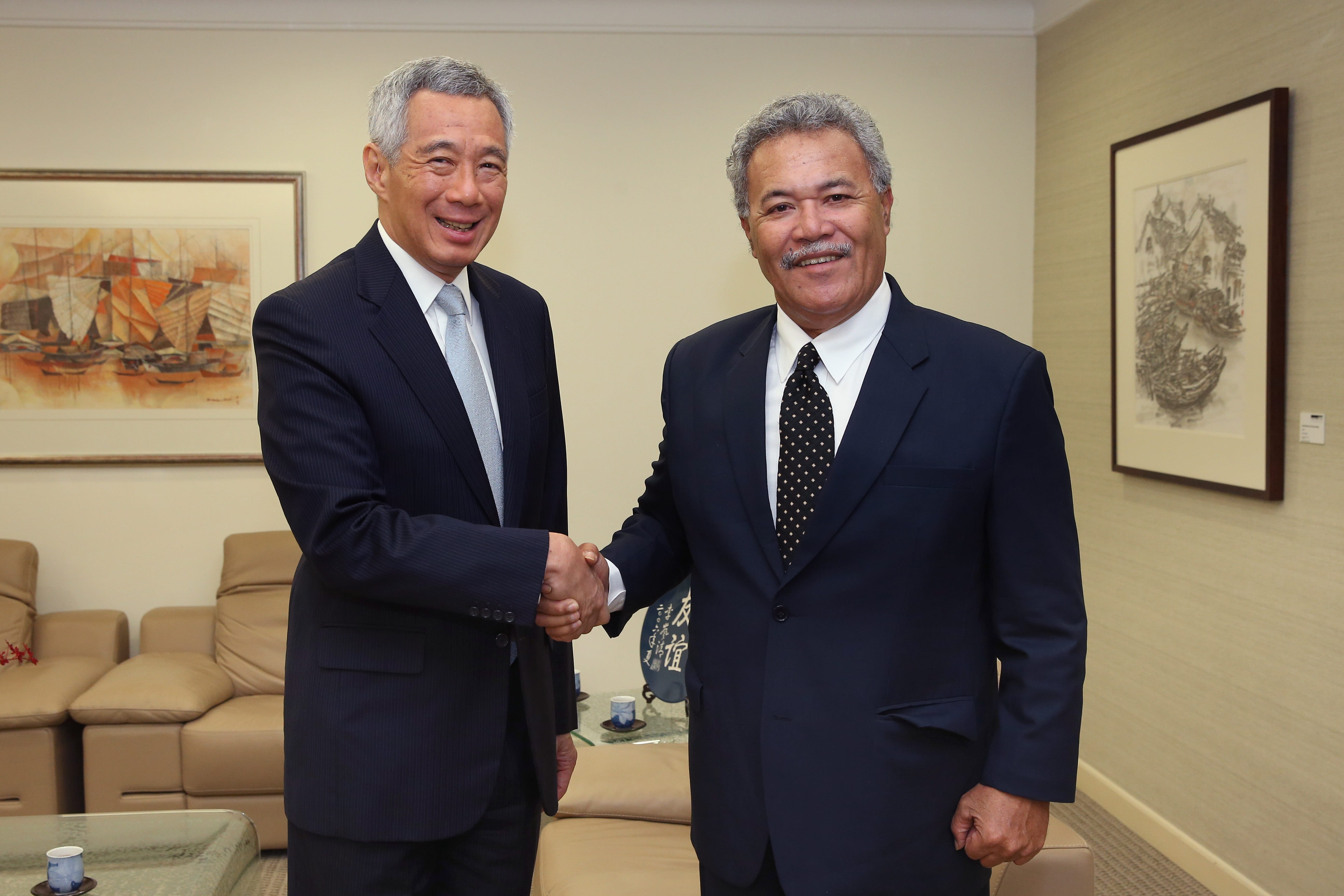 Two men in suits shake hands, smiling, in a room with paintings and furniture.
