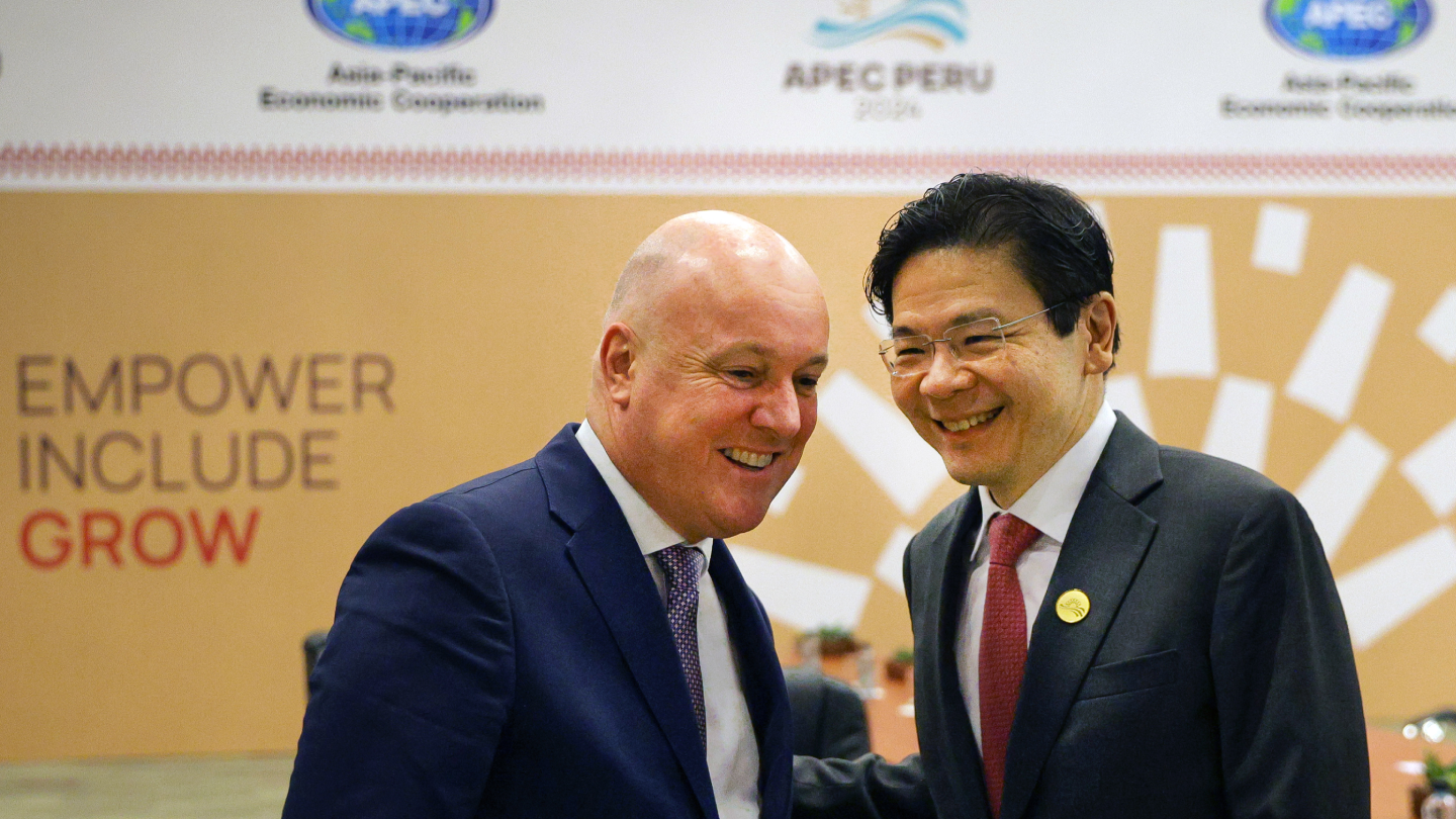 Two men in suits stand close, smiling, against an APEC Peru 2024 backdrop.