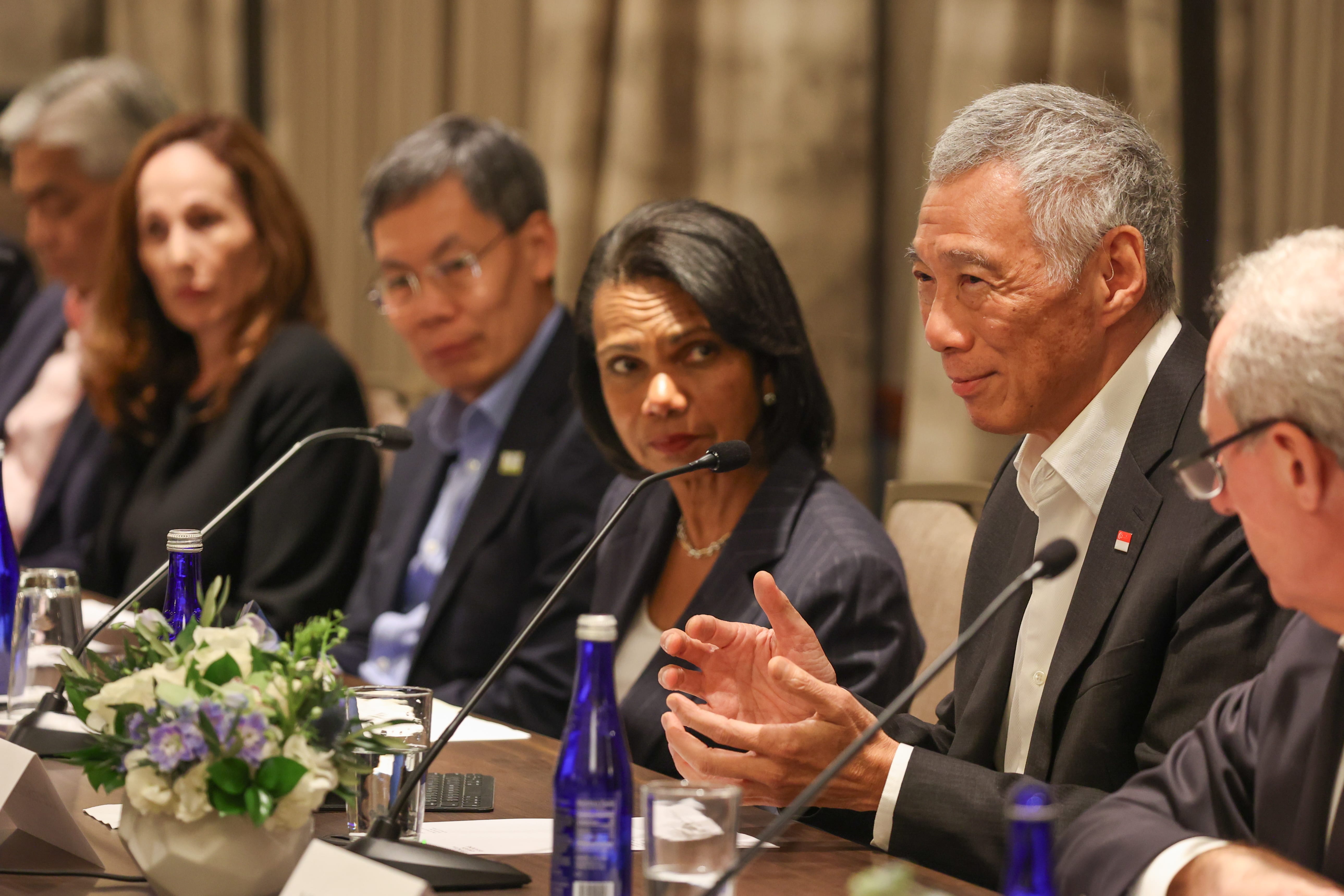 Lee Hsien Loong at a table with others, speaking into a microphone, wearing a suit with a Singapore flag pin.