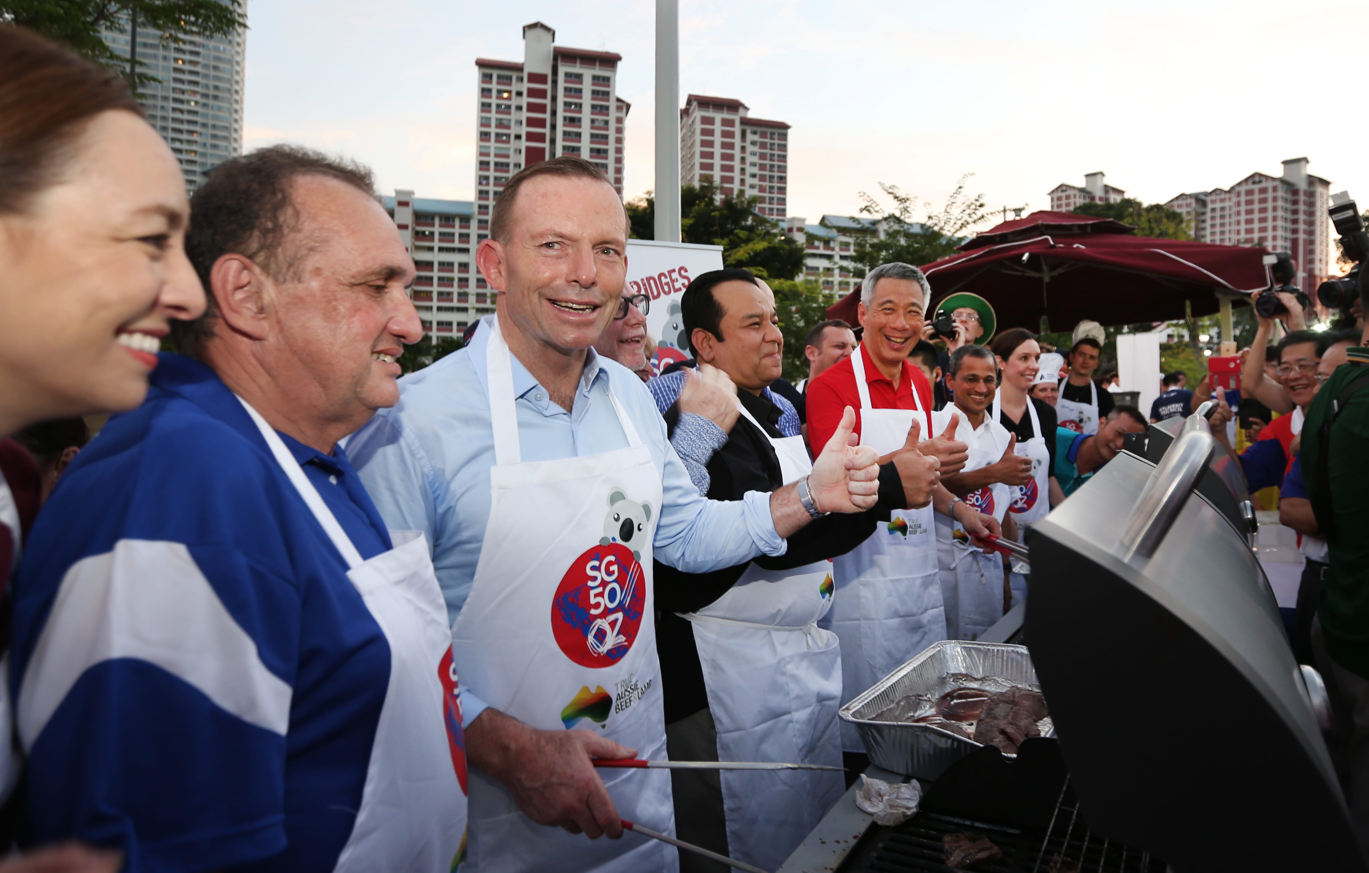 People in aprons grilling meat, thumbs up, urban background. SG50OZ apron design visible.