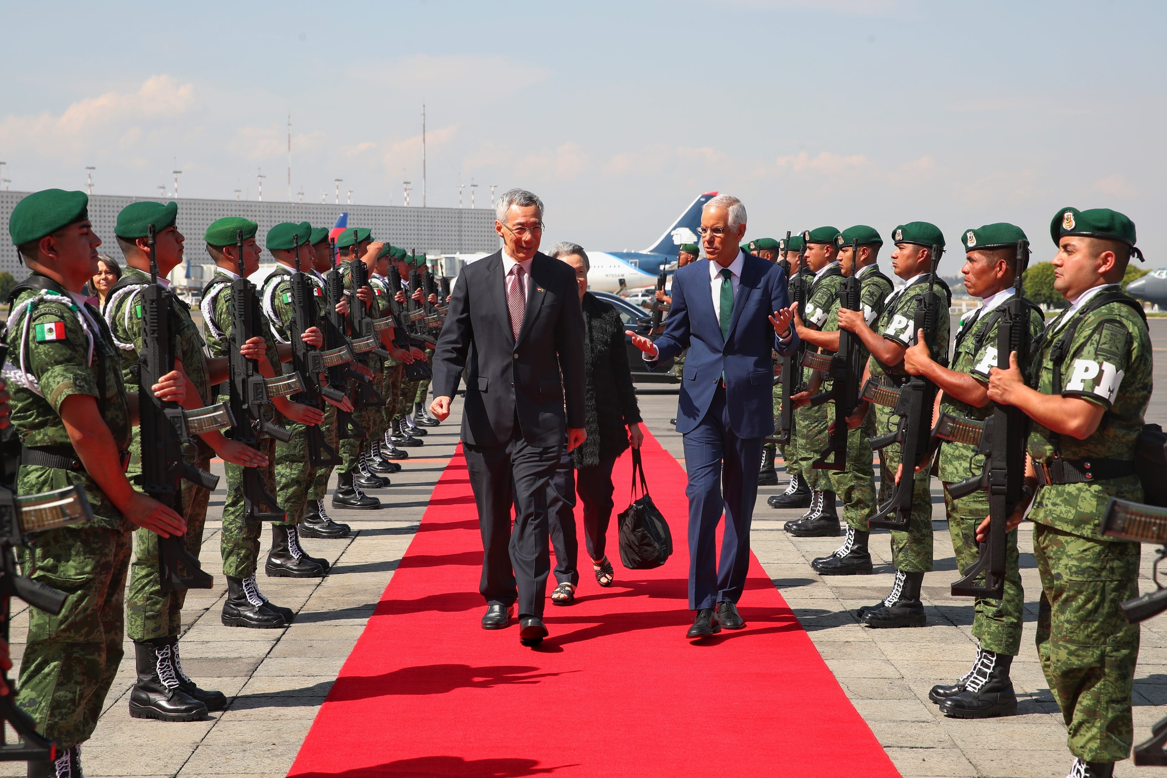 Two men walk on a red carpet flanked by Mexican soldiers holding rifles on an airfield.