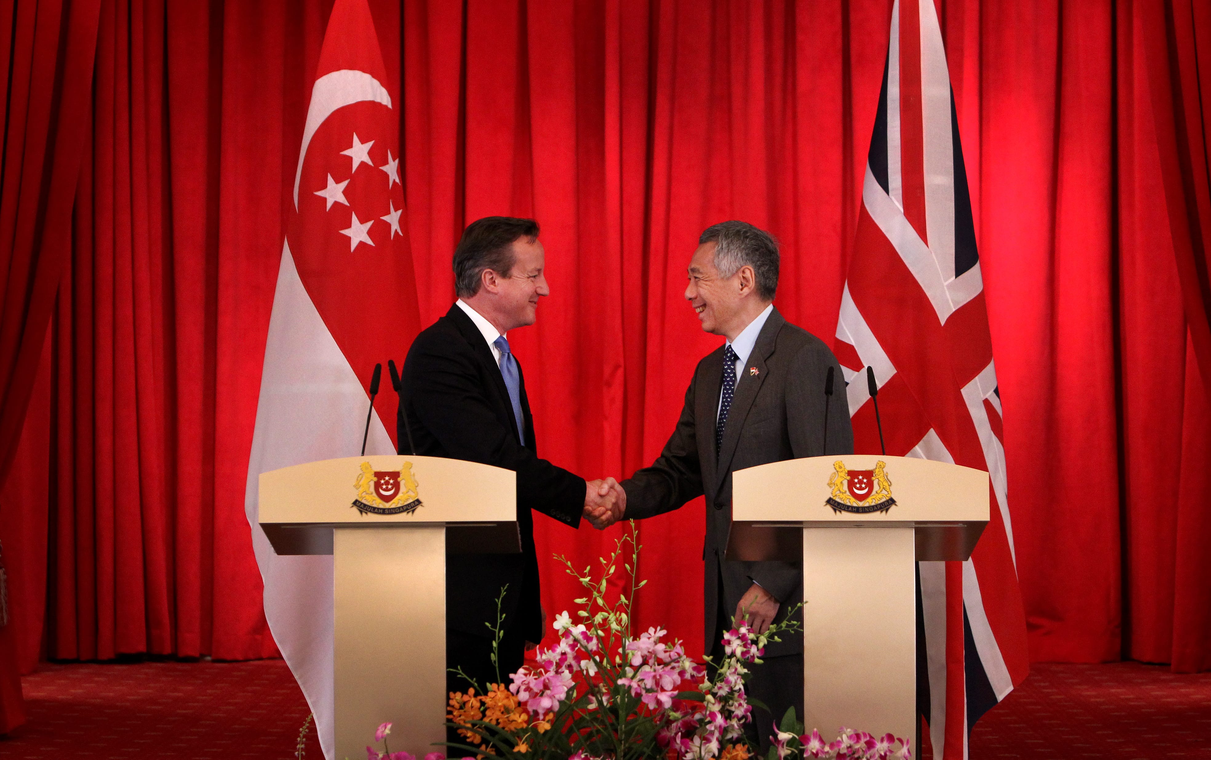 Two men in suits shaking hands before Singapore & UK flags, podiums with heraldry, and flowers.